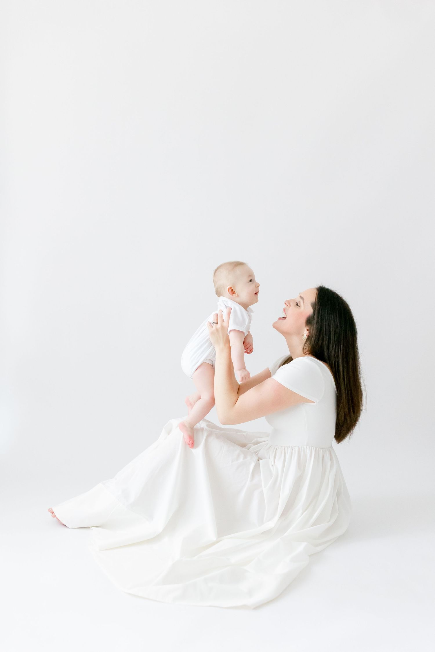 A mother in a white dress sits on the floor holding her baby up, both smiling at each other in a bright studio.