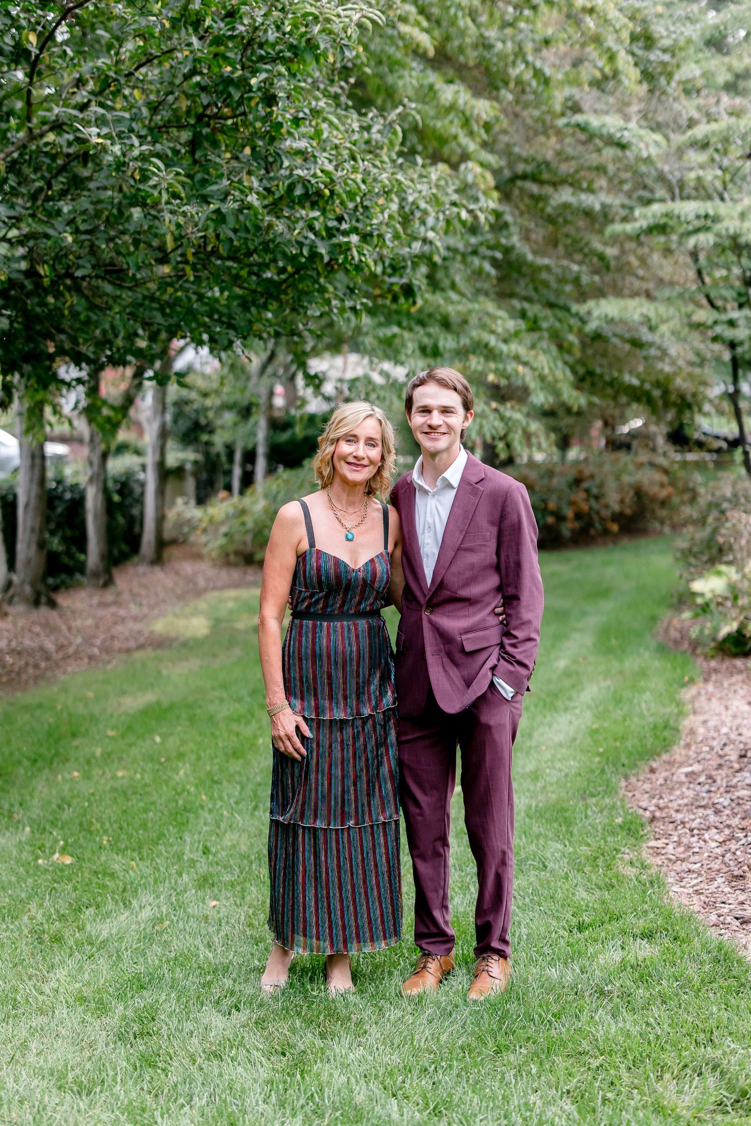 A couple poses together on a grassy outdoor path surrounded by lush greenery and trees.