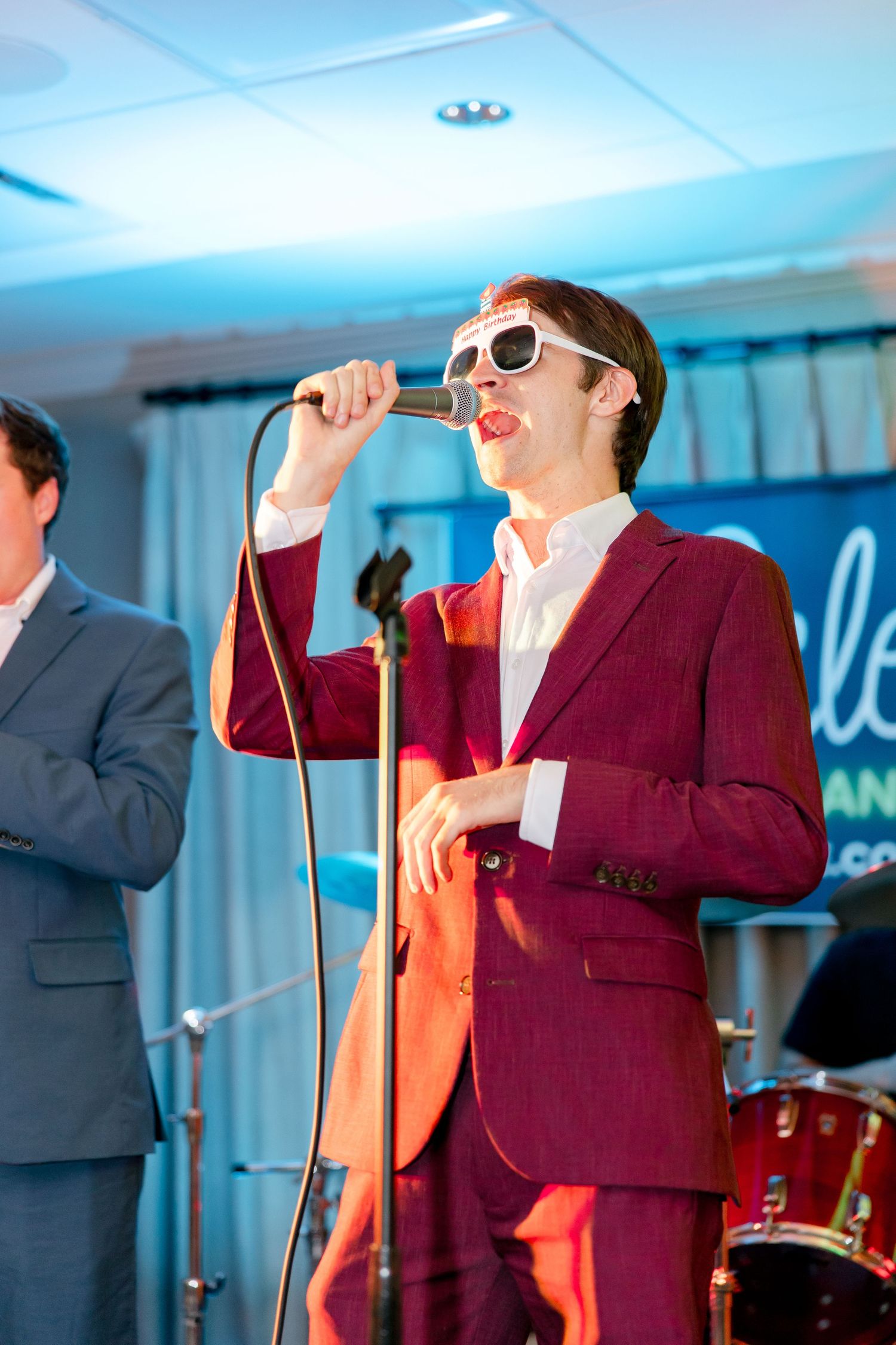 Performer in burgundy suit and sunglasses singing on stage with dramatic blue lighting.