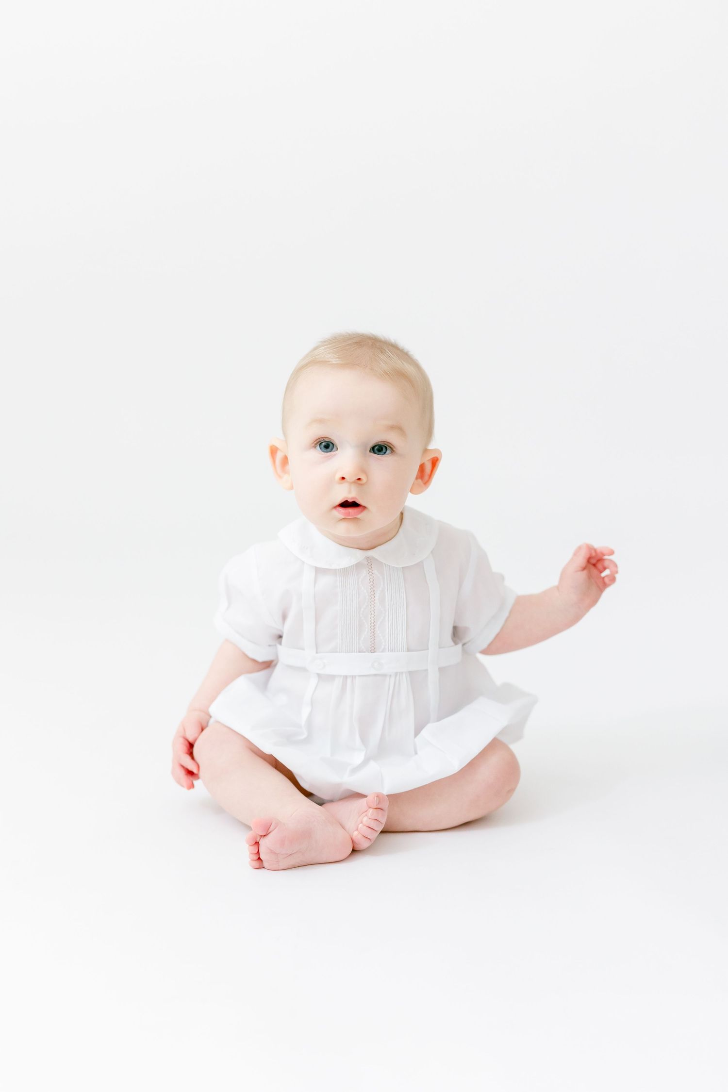Adorable baby in white dress sitting on white background with blue eyes and curious expression.