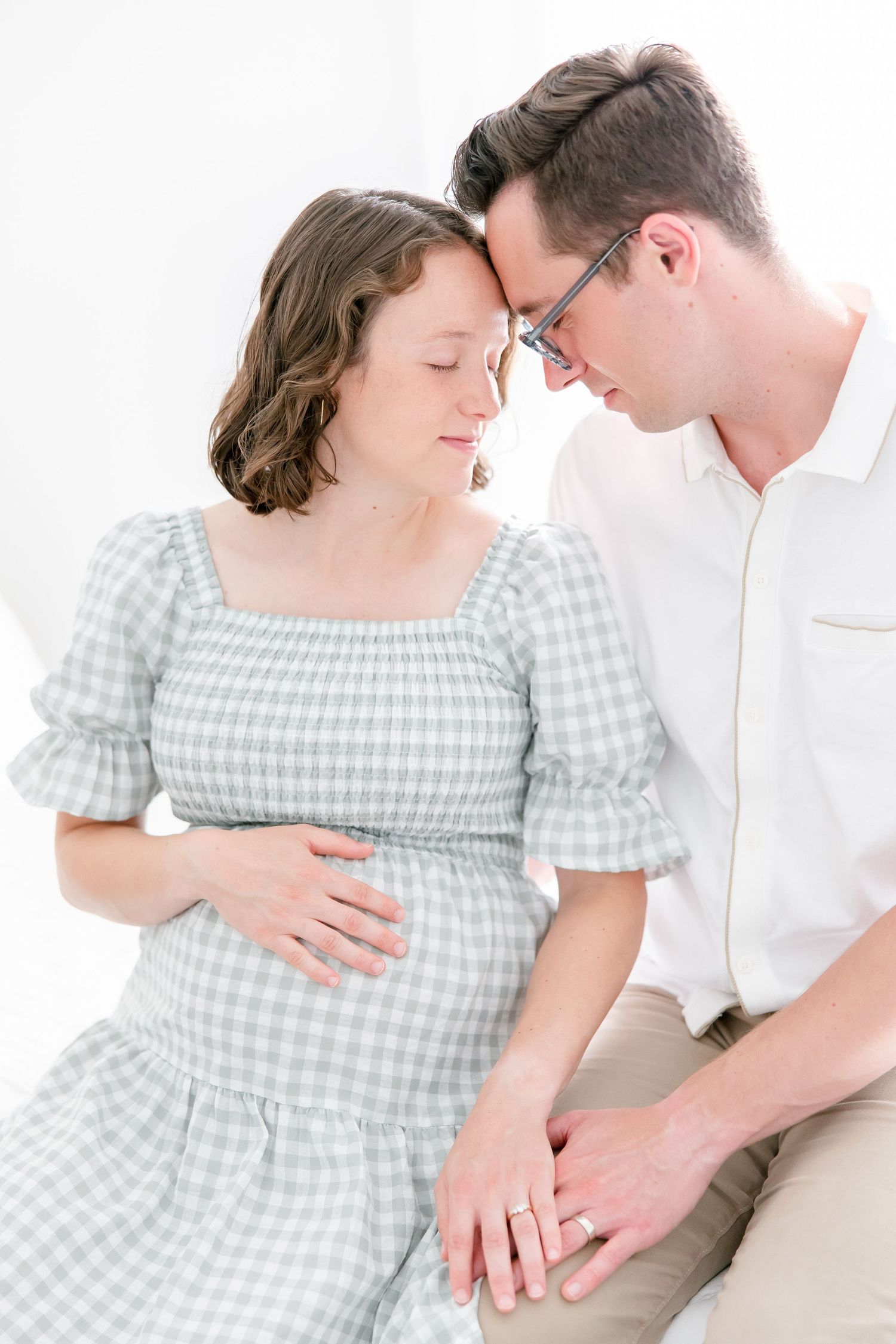 A tender moment between a couple wearing gingham dress and white shirt as they embrace during a maternity photoshoot.