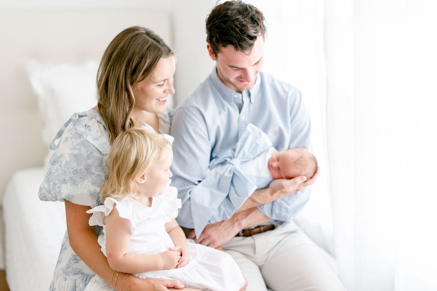 Family sitting together on white couch while looking at newborn baby indoors.
