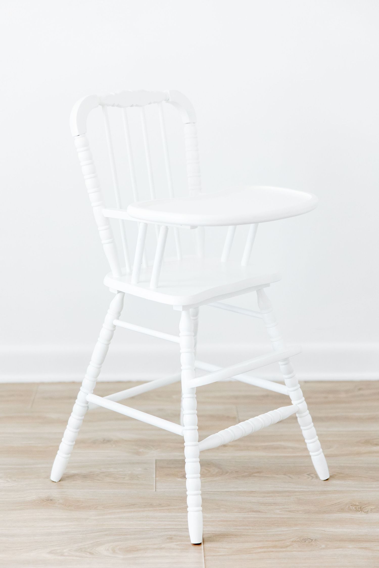 White wooden spindle-back high chair with turned legs on light hardwood floor against white wall.