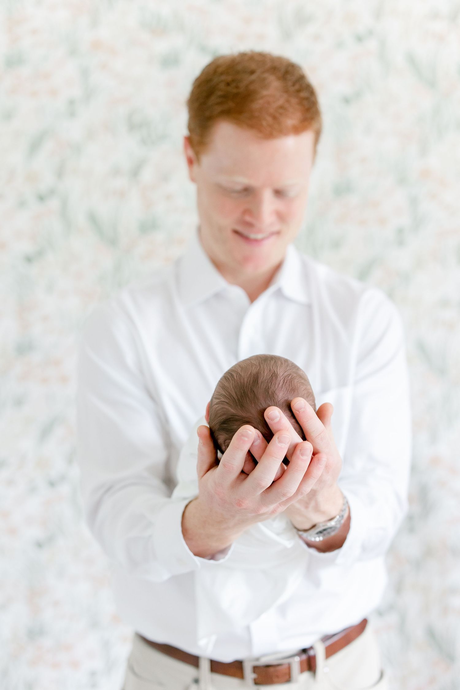 A person in white shirt holds sleeping newborn against floral wallpaper.