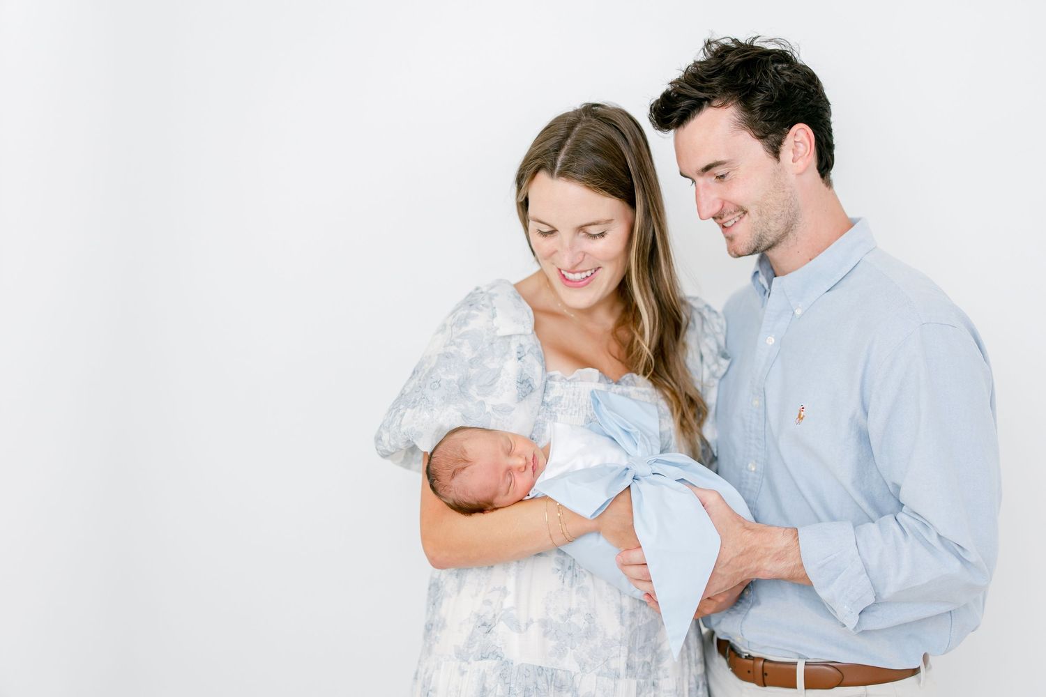 A couple in soft blue and white attire lovingly gazes at their newborn baby against a white backdrop.