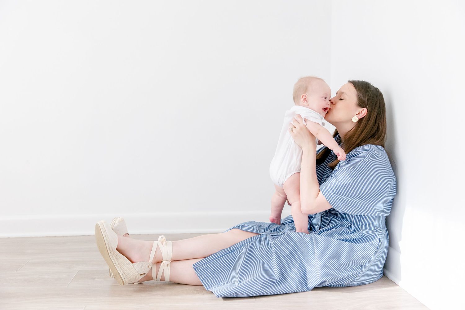 Parent in blue dress sits on floor playing with baby in white outfit against neutral background.