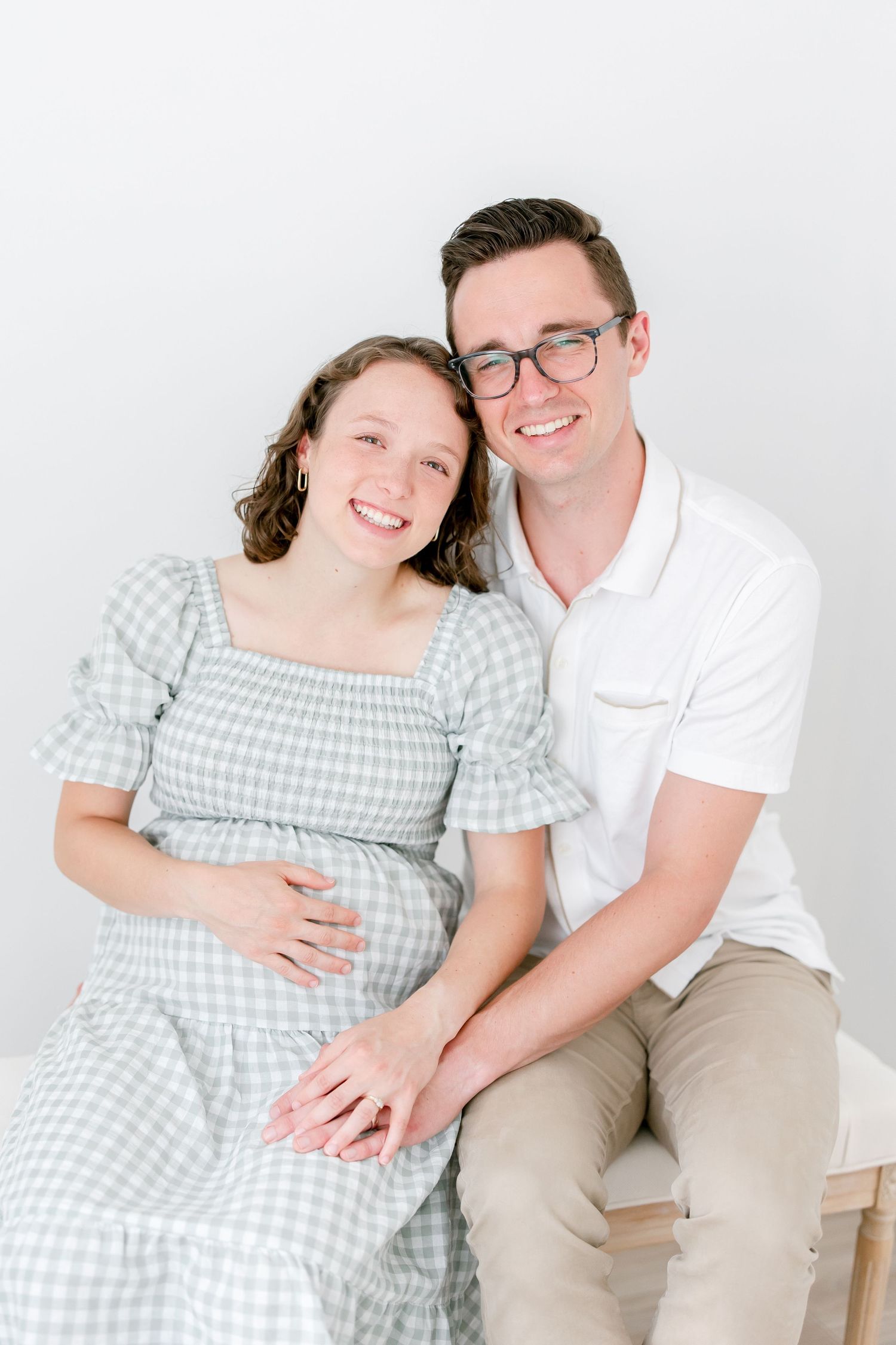 A couple in white and gingham sits together for a maternity photo against a light background.