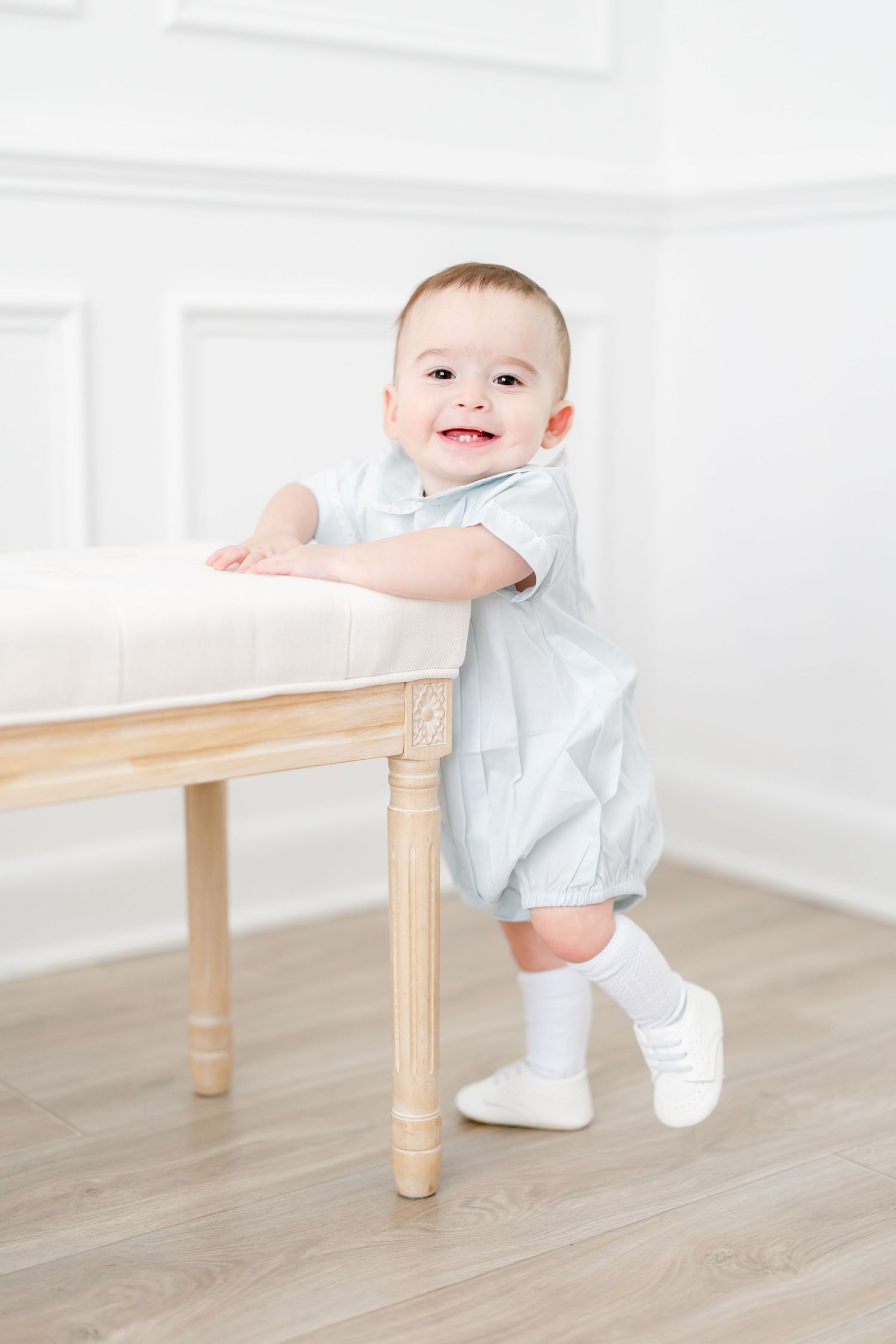Baby in white outfit holding onto wooden furniture while standing on light hardwood floor in bright room.