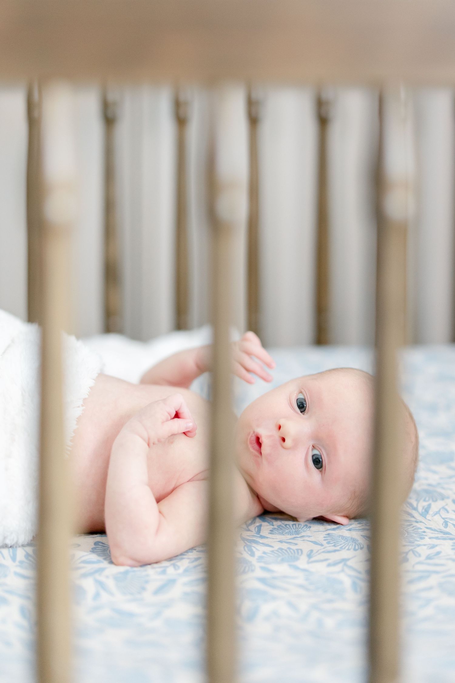 Baby lying in white crib on light blue bedding viewed through wooden slats.