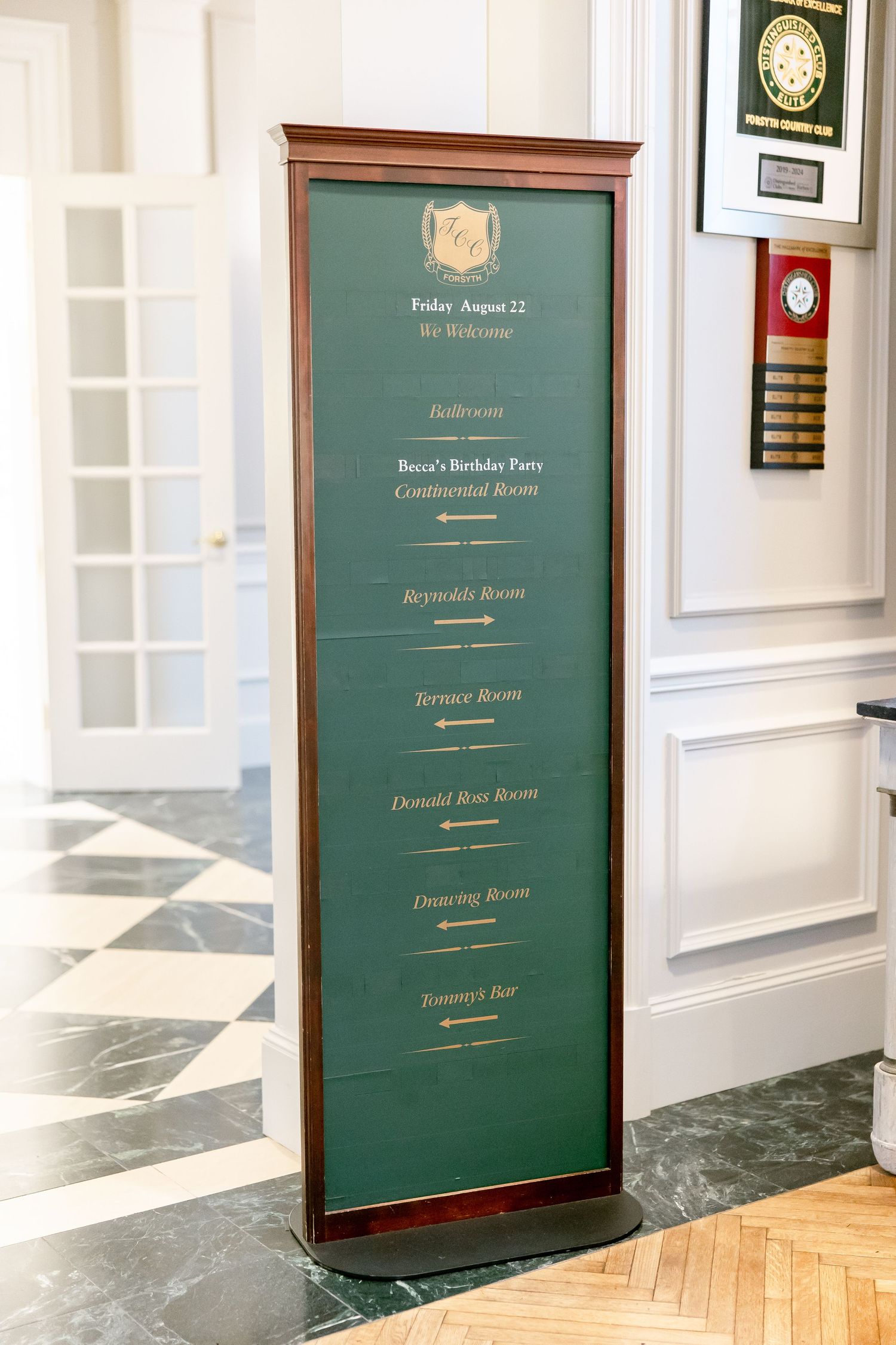 Elegant green and gold event directional signage displayed in wooden frames along a white hallway.