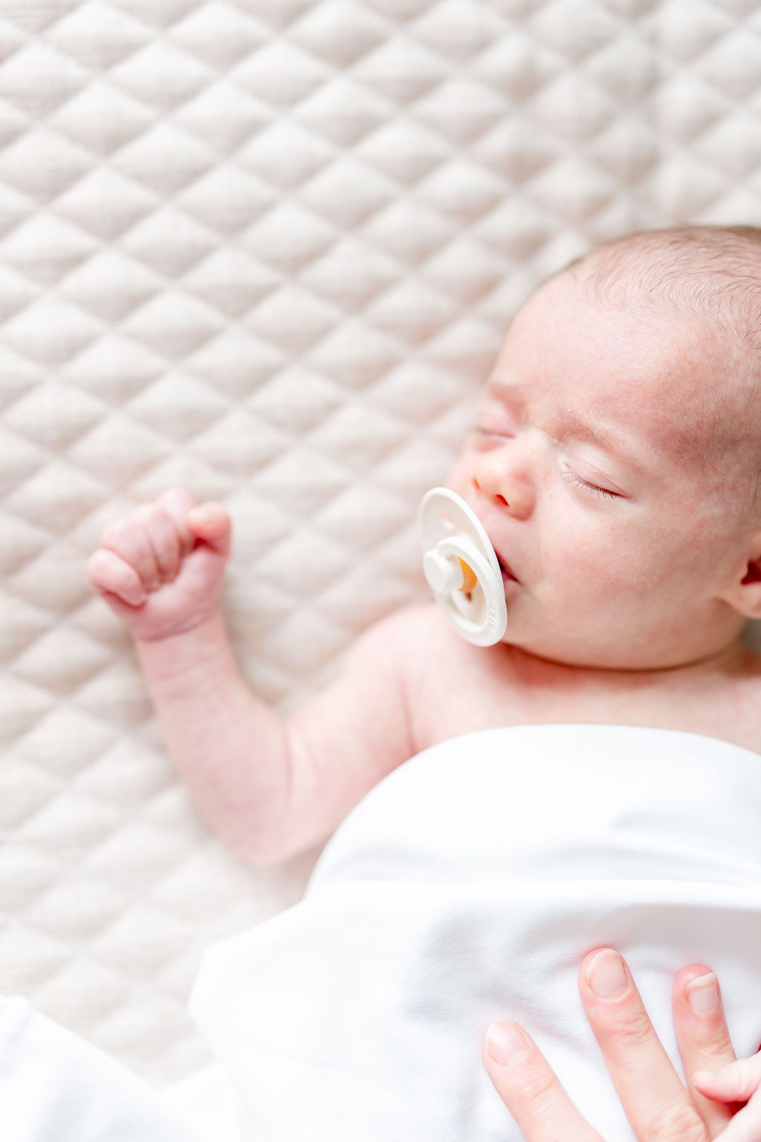 Sleeping newborn baby wrapped in white blanket against textured white background.