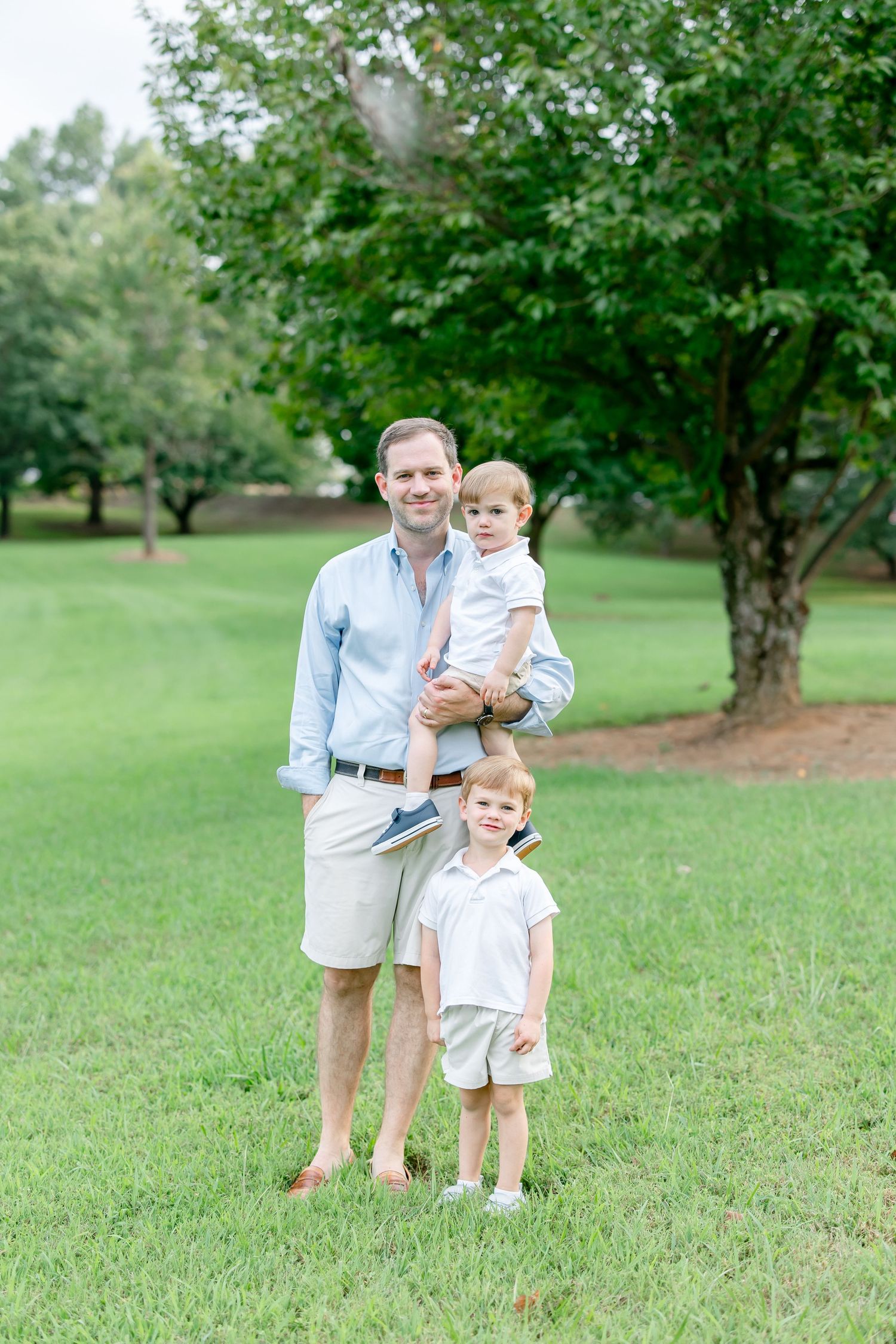 Family members in coordinating light blue and white outfits pose together on grassy lawn in outdoor park setting.