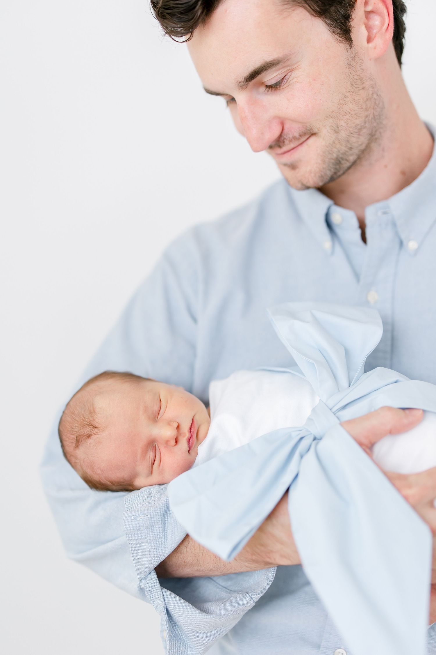 A newborn baby sleeps peacefully in white swaddle while being cradled in someone wearing a light blue shirt.