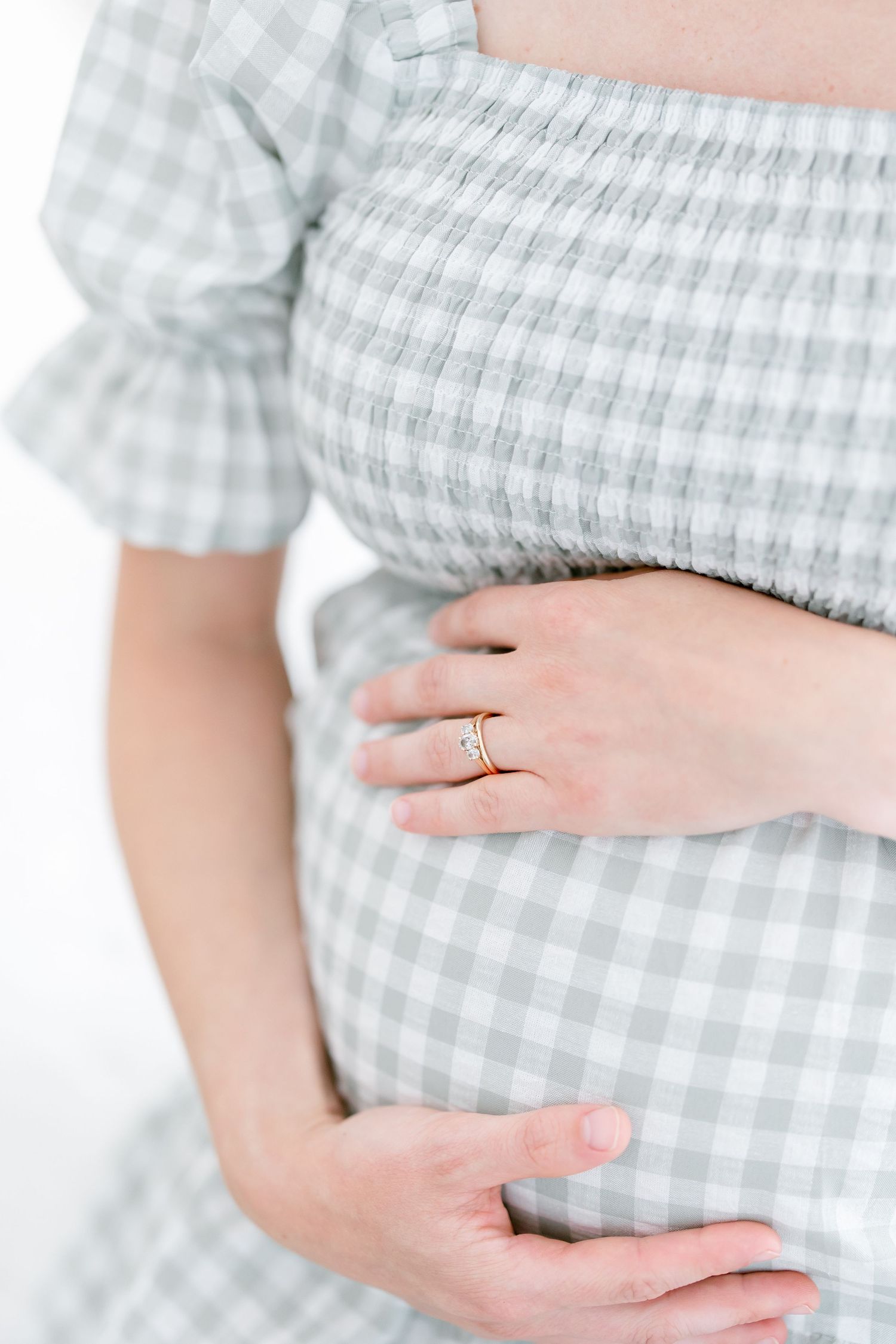Hands gently cradling a pregnant belly in a soft gray and white gingham dress.