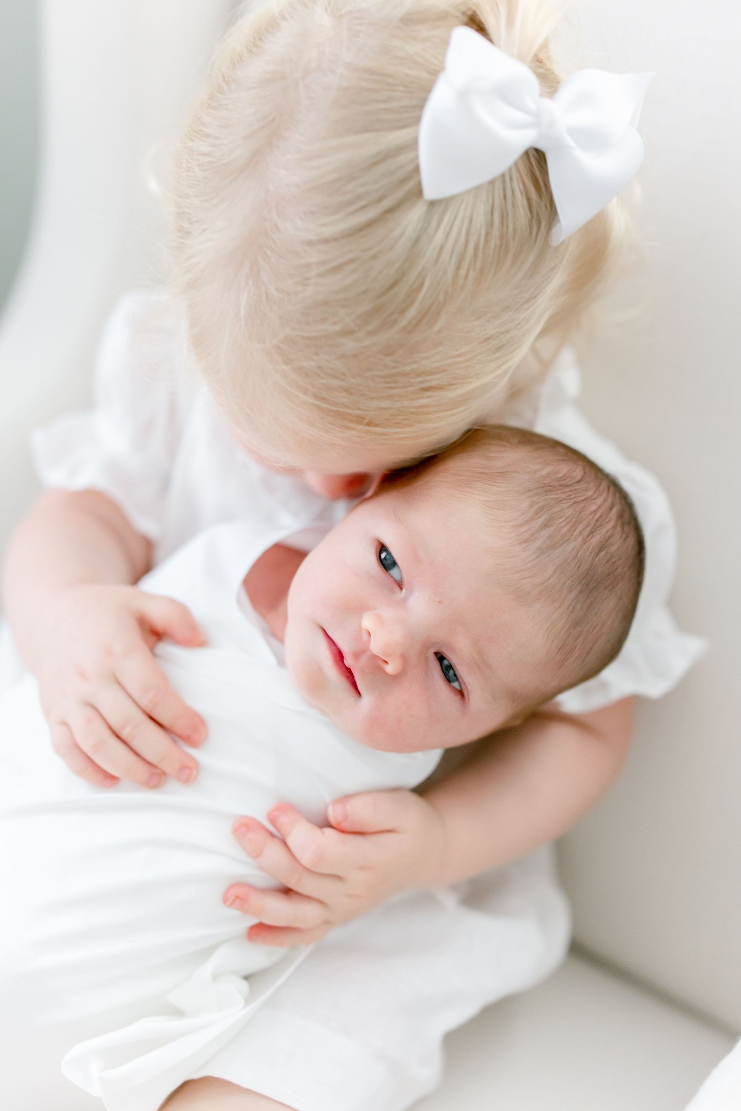 Sweet moment between siblings as toddler in white dress with bow hugs newborn baby.