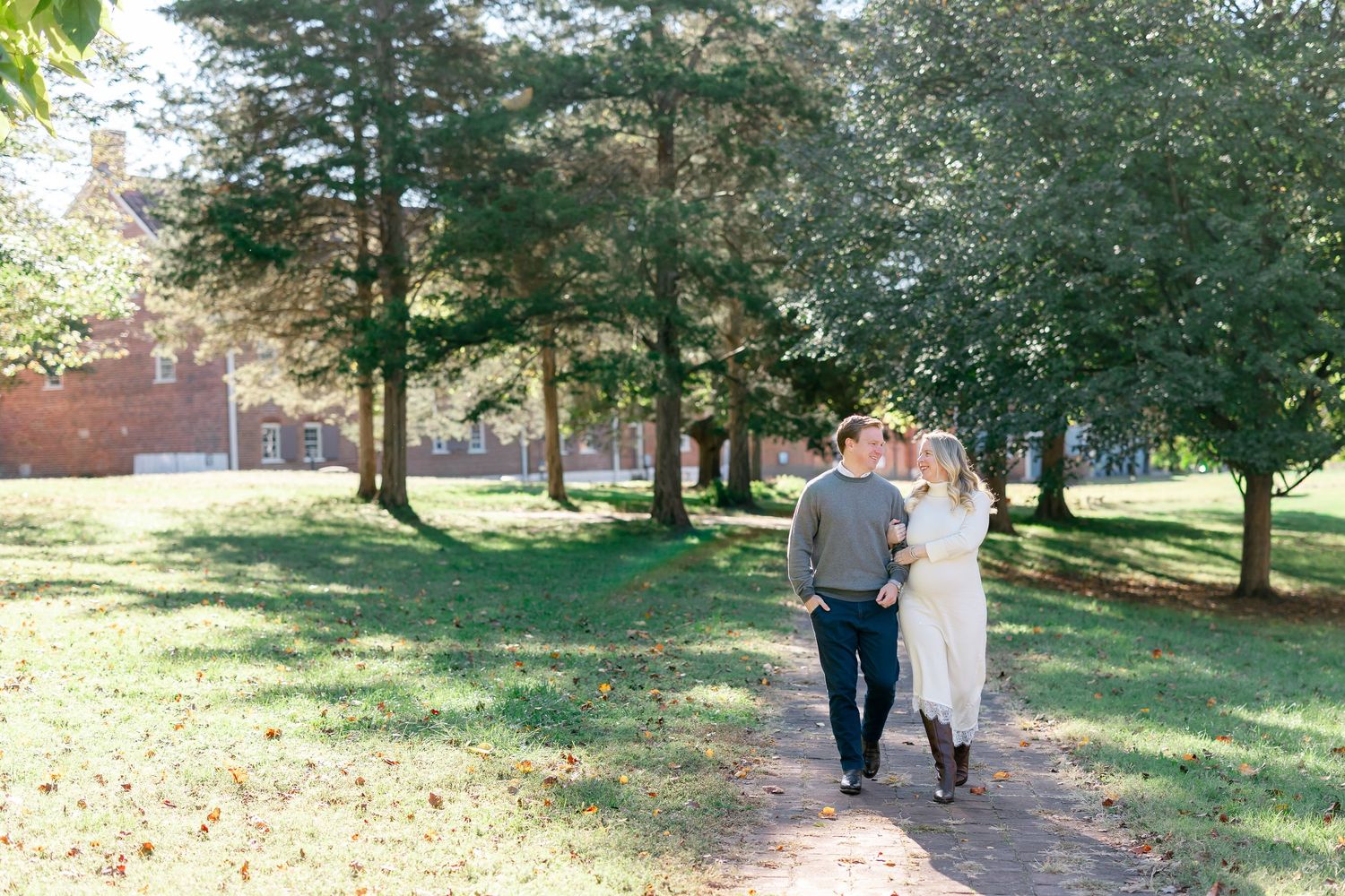 Couple walking arm in arm down a sunlit dirt path lined with tall trees and green grass, with a red brick building in the background.