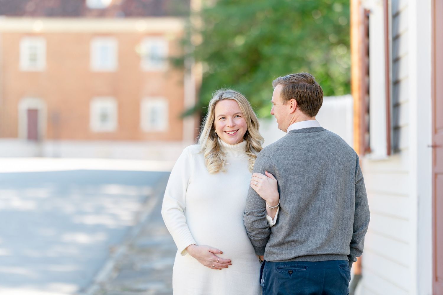 Pregnant woman in a white sweater dress cradles her belly as her partner in a gray sweater embraces her on a sunlit historic brick street.