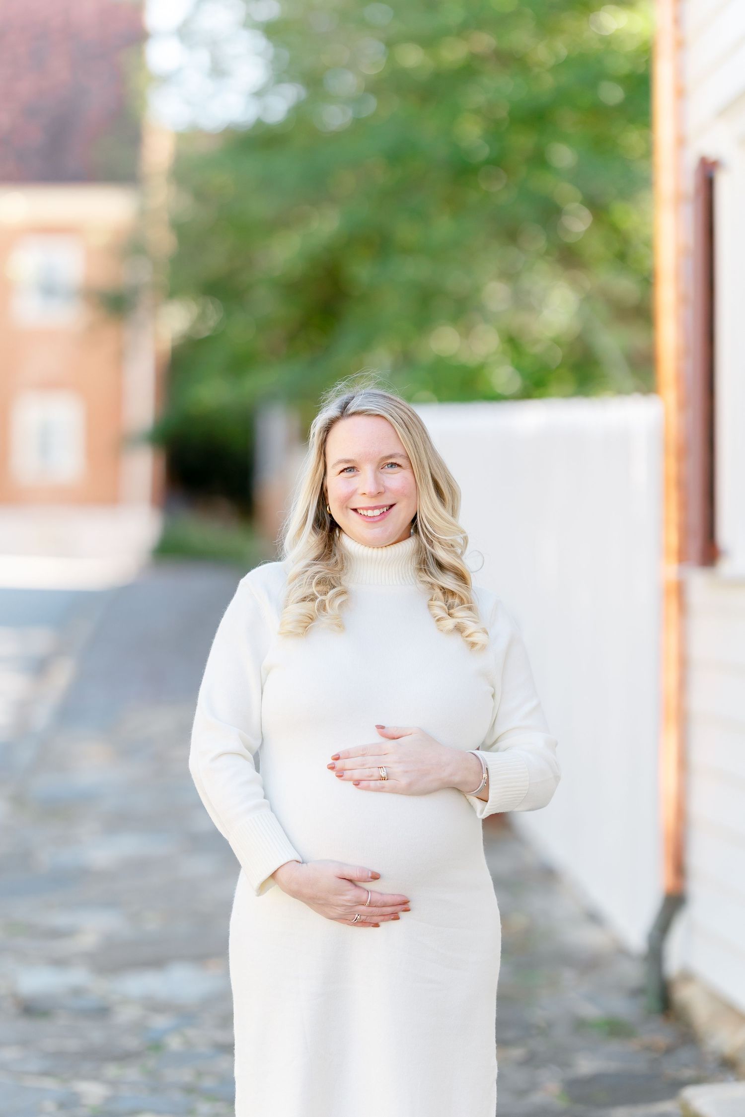 Smiling pregnant woman in a cream turtleneck dress cradles her baby bump on a sunlit cobblestone street lined with historic buildings.