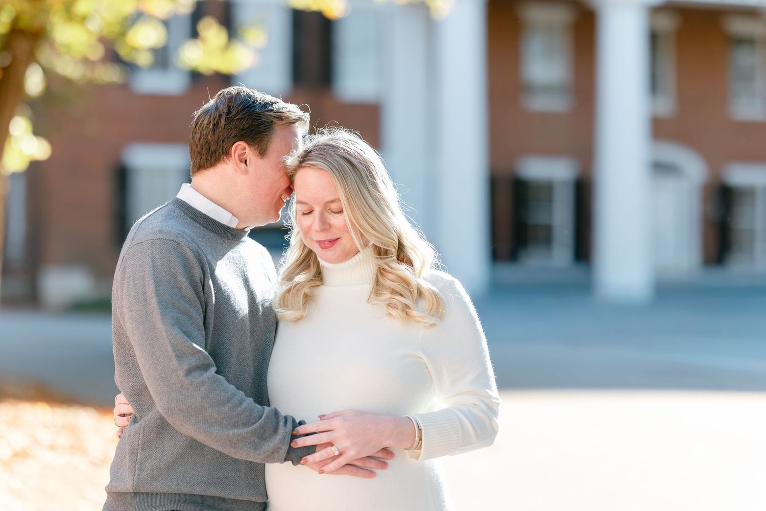 Couple embracing outdoors as a man kisses a blonde woman's forehead in front of a brick building with white columns in autumn.