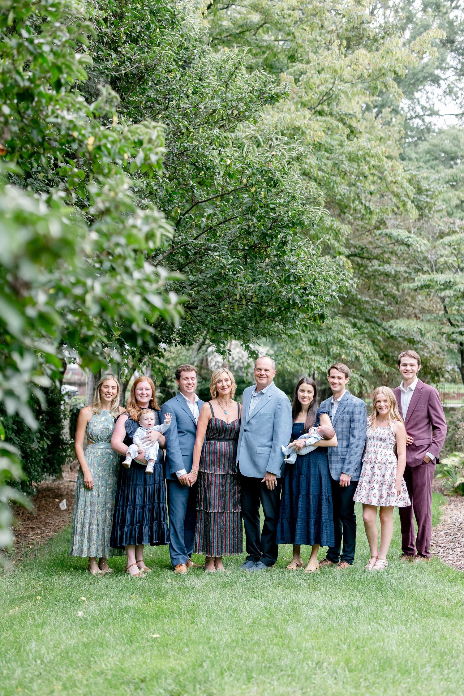 Large family group portrait posing outdoors in a lush garden, dressed in elegant formal attire for a special occasion.