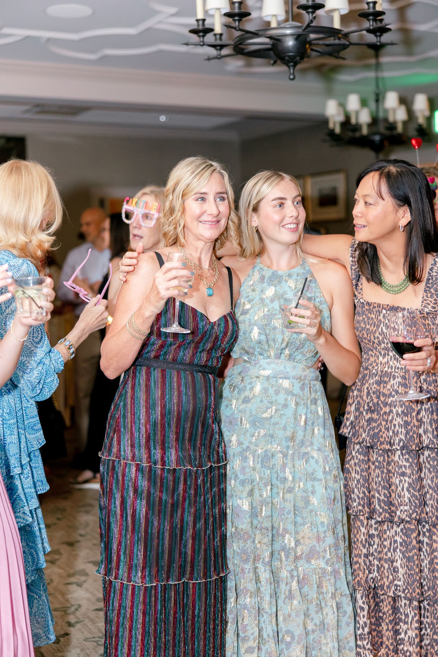 A group of women in patterned dresses enjoying a celebration indoors with drinks and laughter.