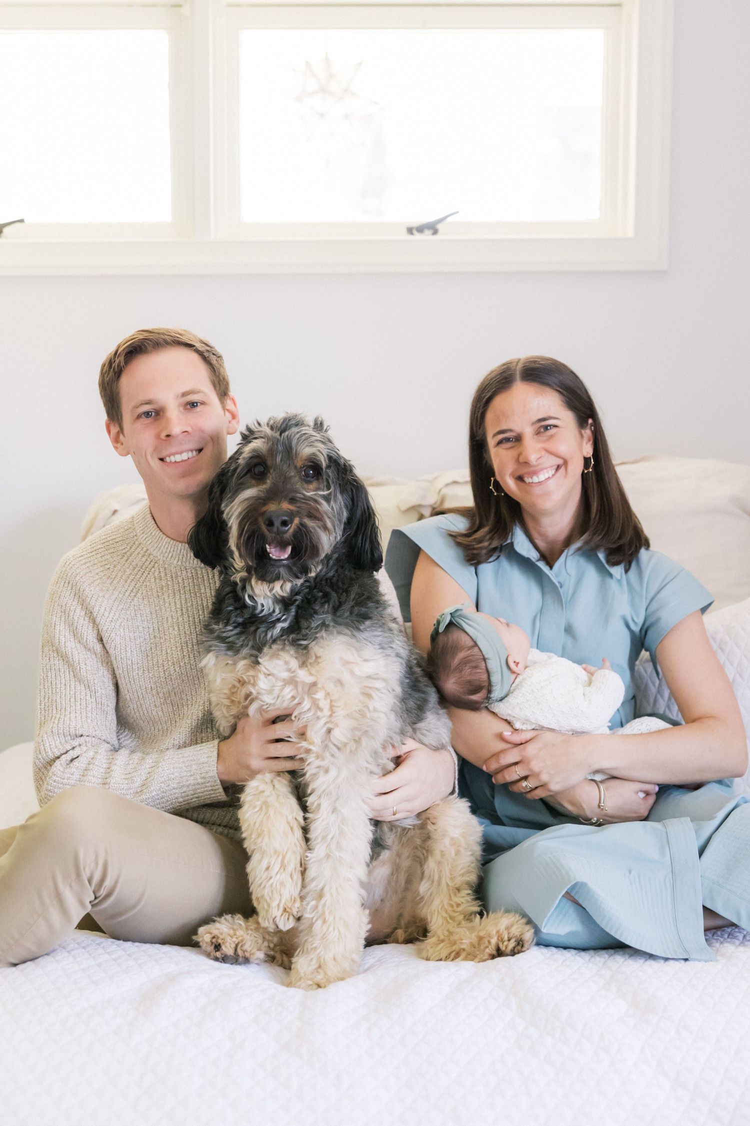 Family pet photography series showing a fluffy dog lounging with owners on white bedding in bright natural light.