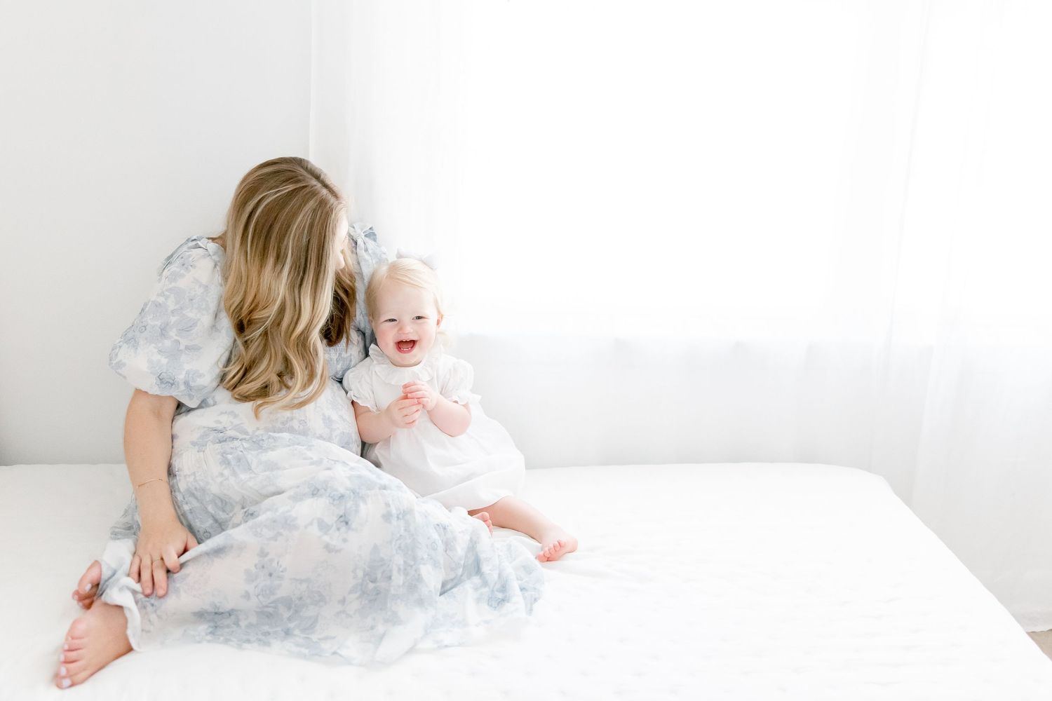 A baby and mom in a white dress cuddle on a light gray bed with white bedding and blue accents.