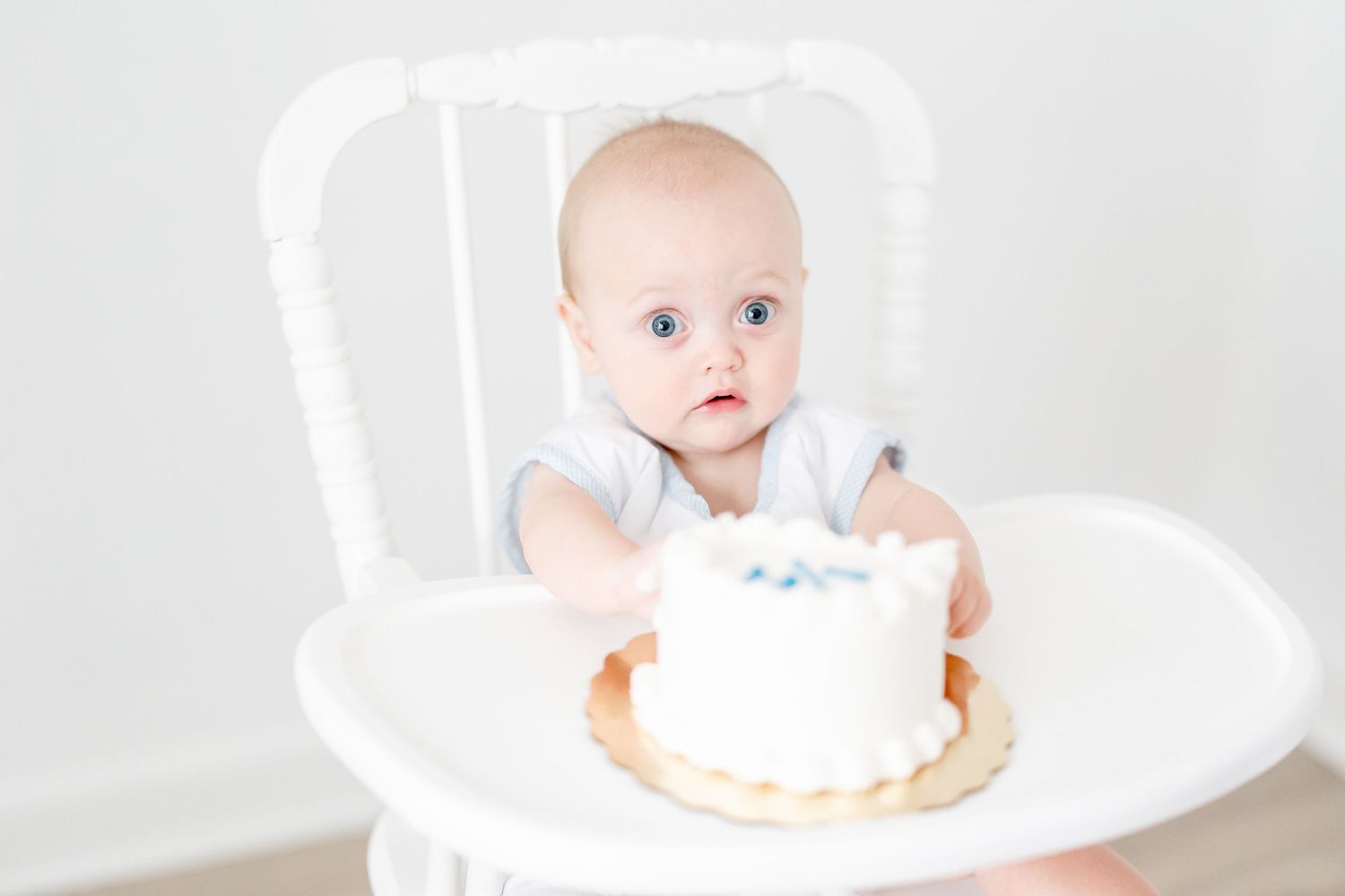 Baby seated in white high chair with birthday cake for cake smash photography session.