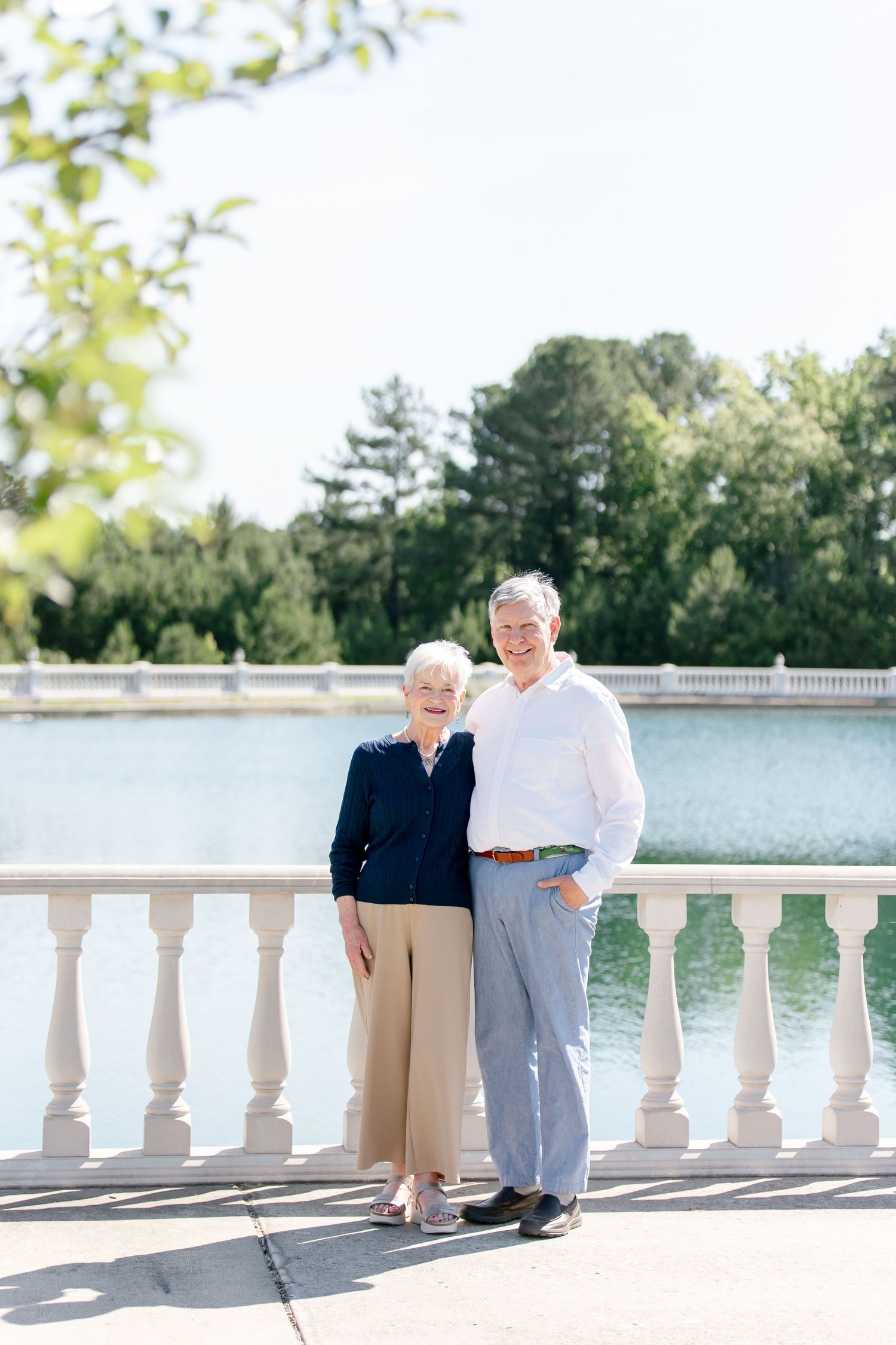 Two people in light clothing pose lovingly by a white balustrade overlooking a serene lake on a sunny day.