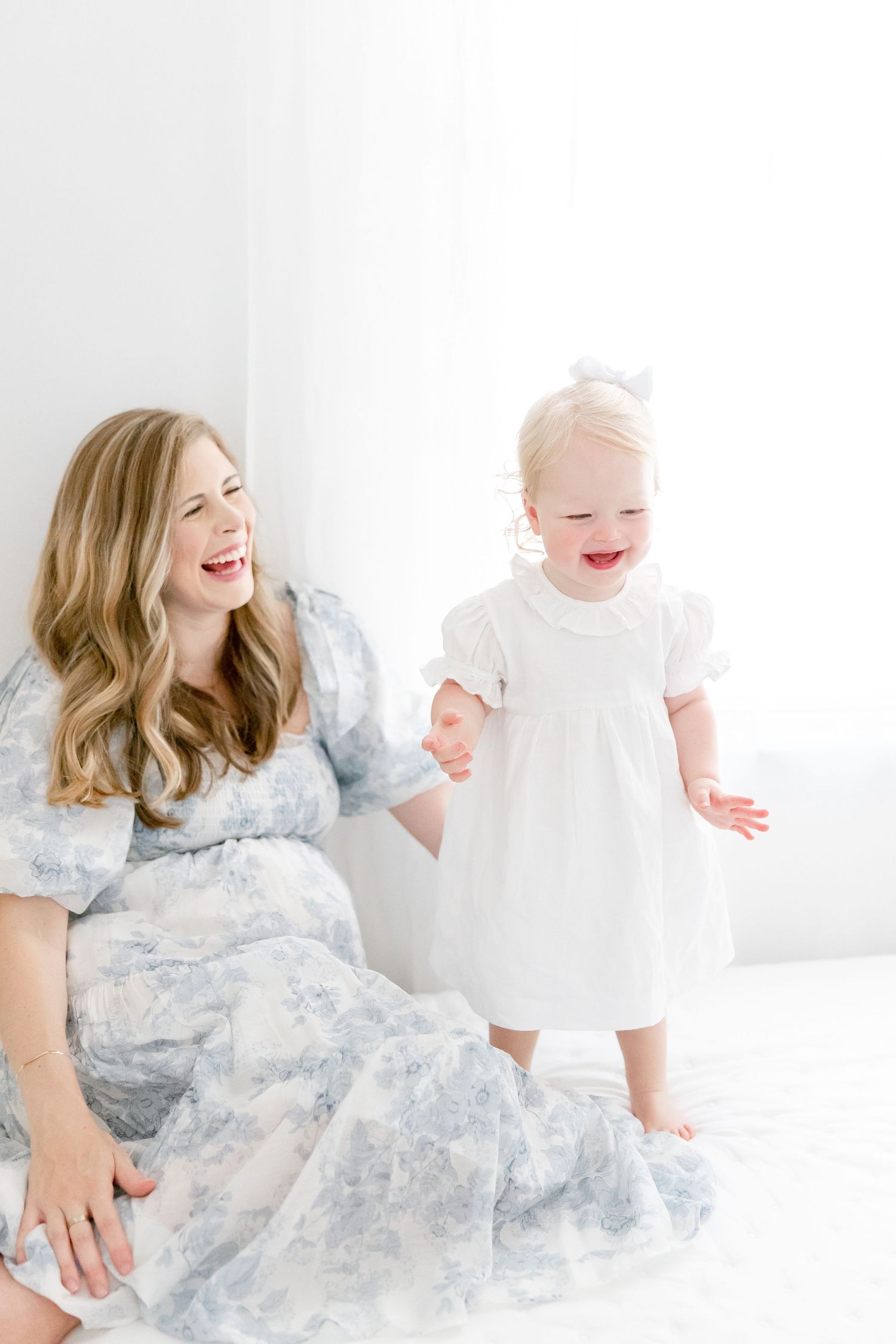Two people wearing light blue and white dresses share laughter in a bright white studio setting.