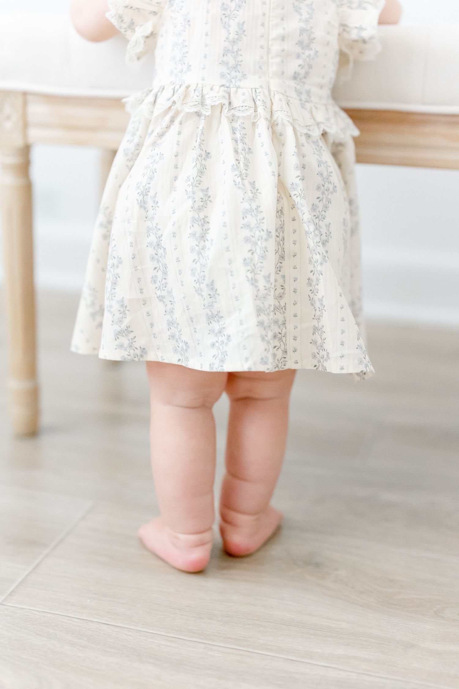 Close-up of tiny bare feet wearing a delicate white floral dress with ruffles on light hardwood flooring.
