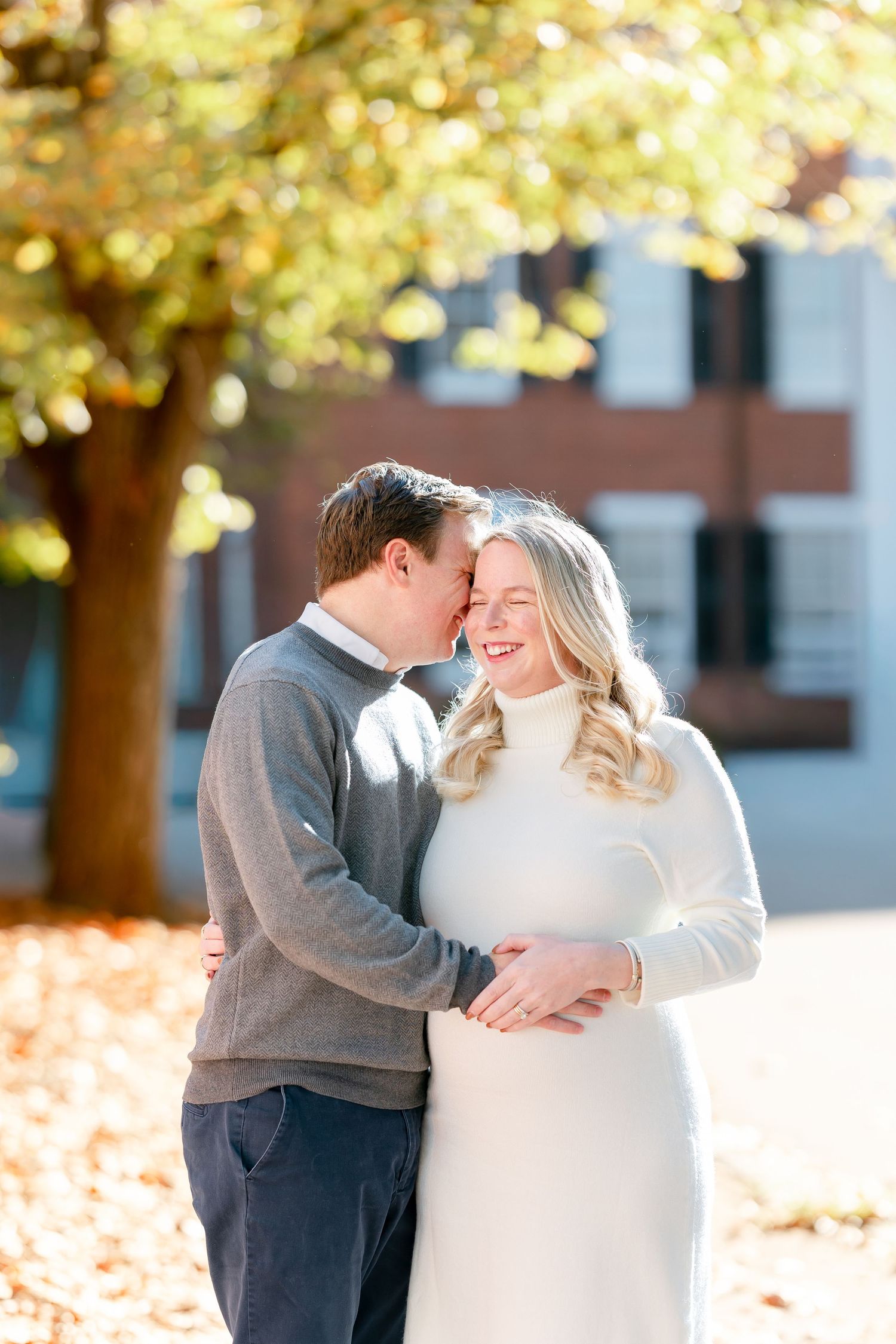 A smiling couple embraces outdoors on a sunny autumn day, the man kissing the woman's forehead near a brick building and golden fall leaves.