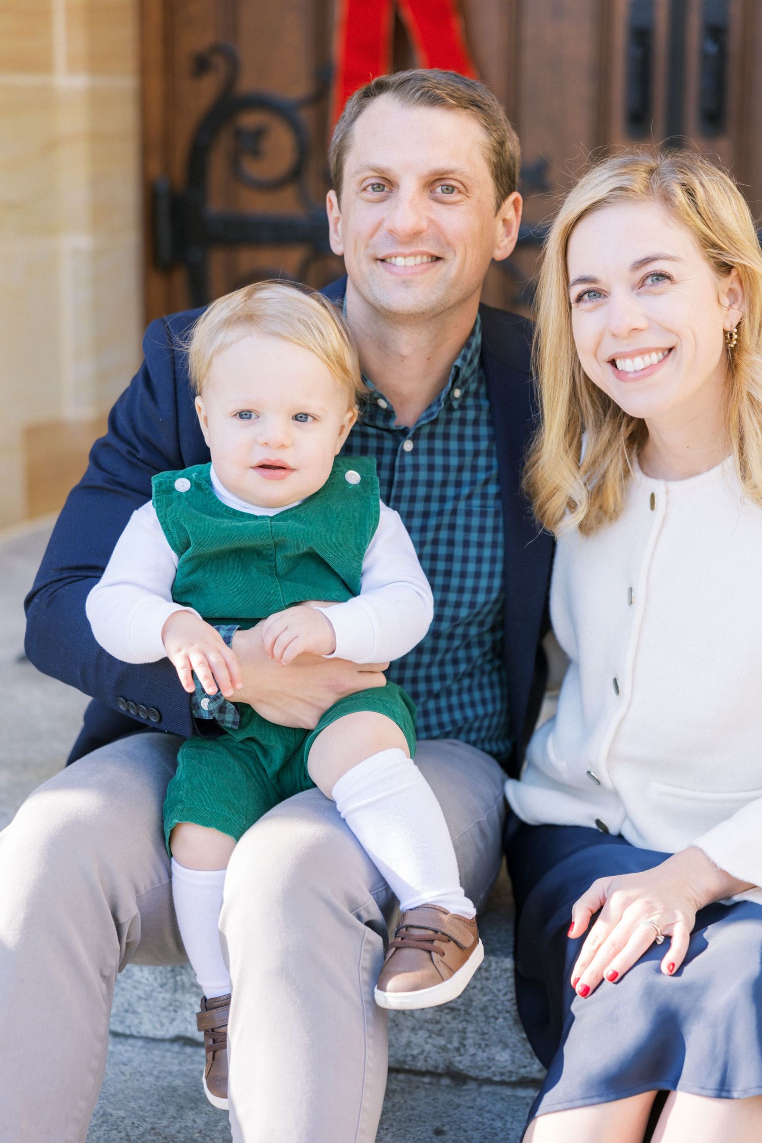 Family sitting on outdoor steps with small child wearing green overalls and white dress shirt.