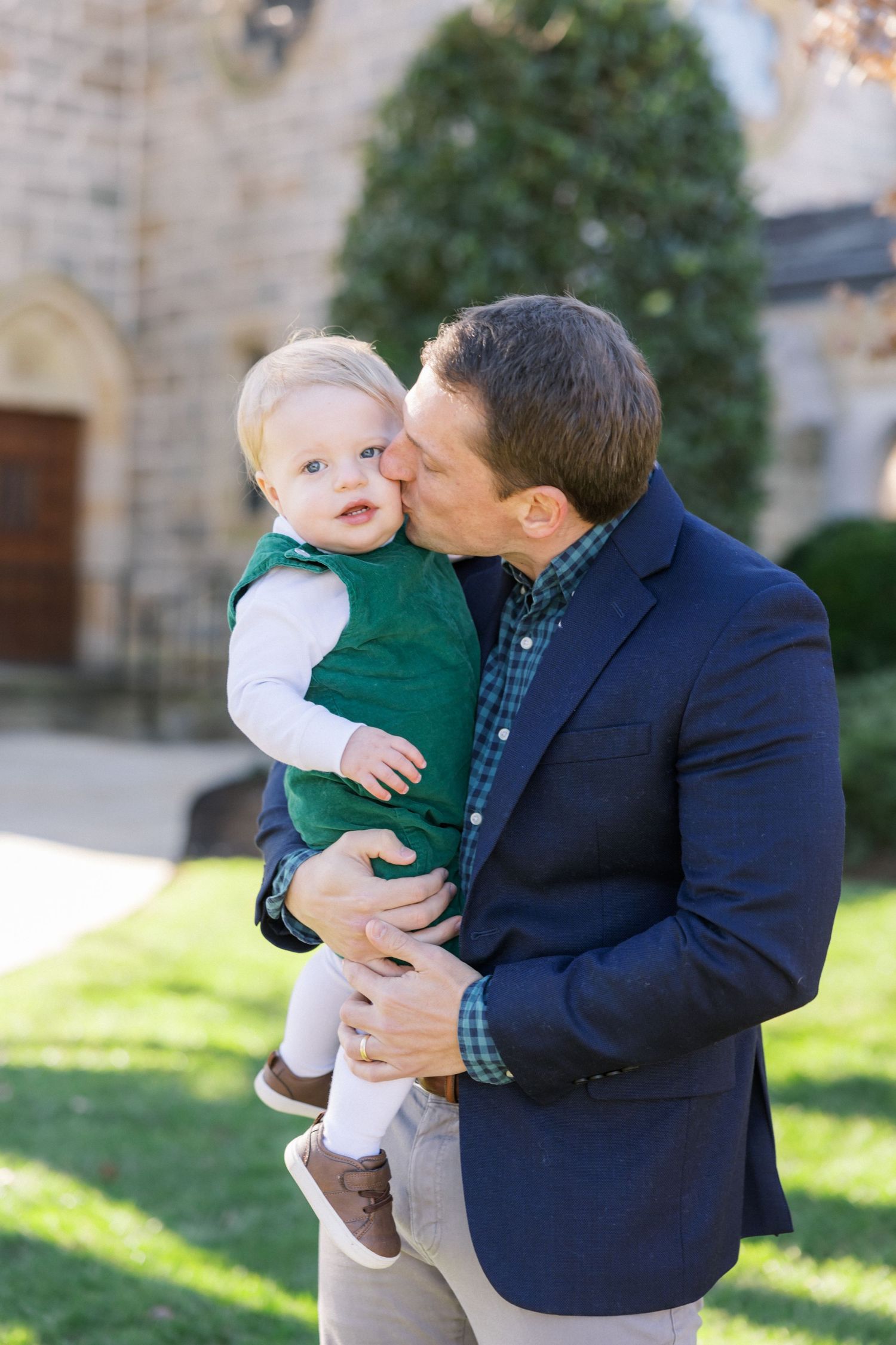 A parent wearing a navy blazer kisses a small child in a green vest outside a stone church.