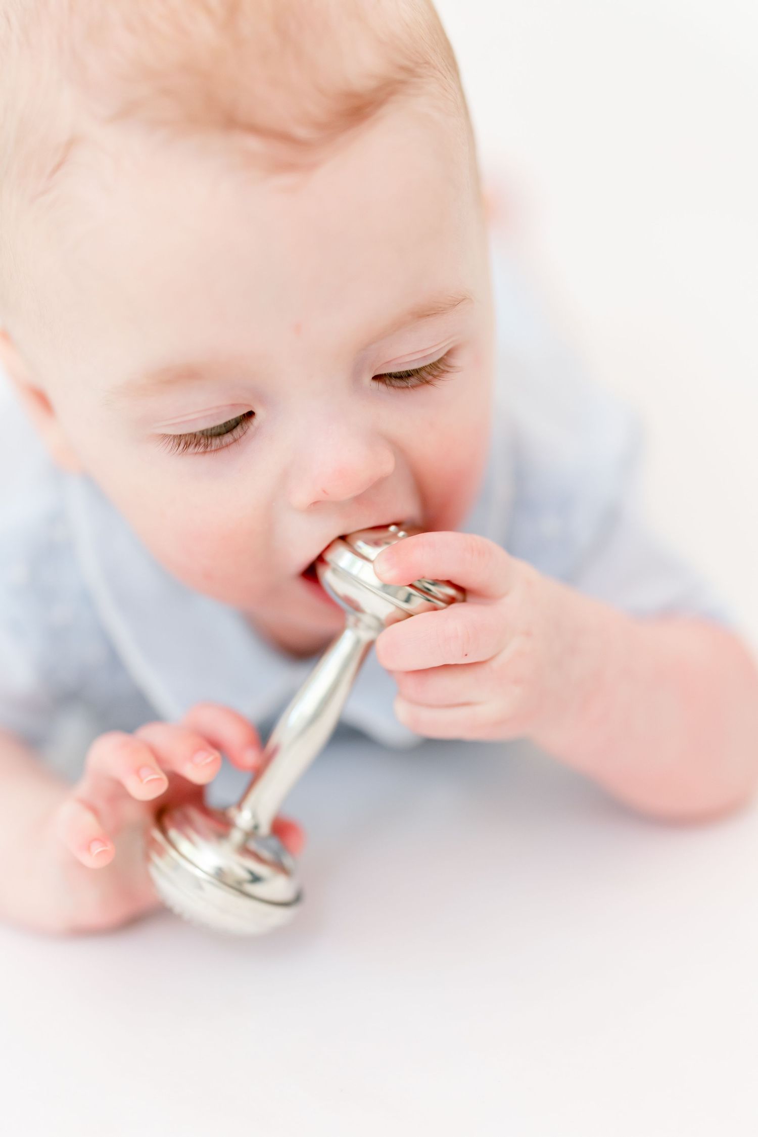 Baby in a light blue outfit lying on their tummy and chewing on a silver dumbbell-shaped rattle against a white background.