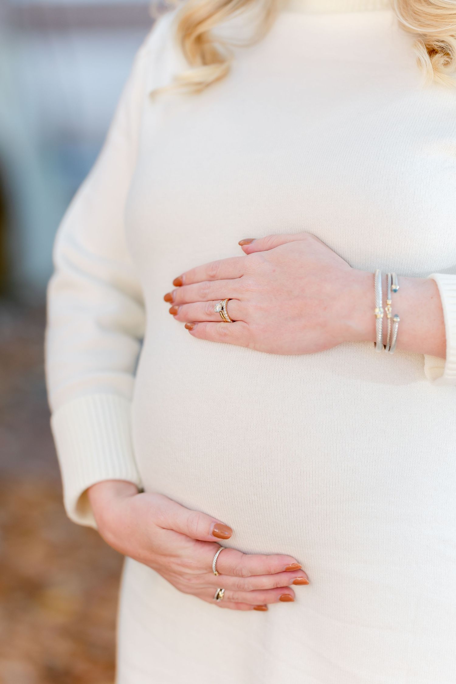 Pregnant woman in a cream knit sweater dress cradling her baby bump with both hands, wearing rings and silver bracelets.