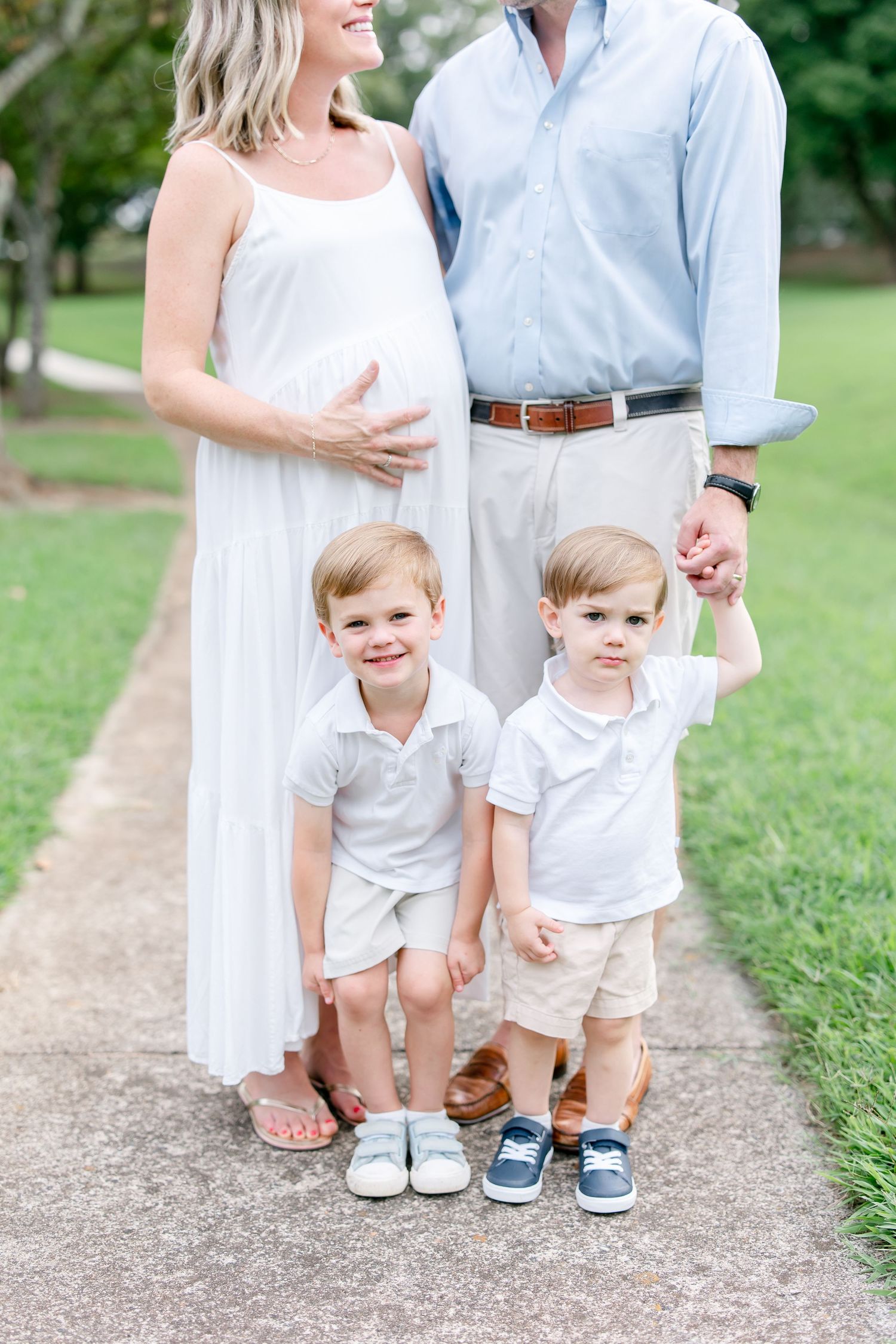 Two small children stand together in white outfits on a gravel path while their parents look on.