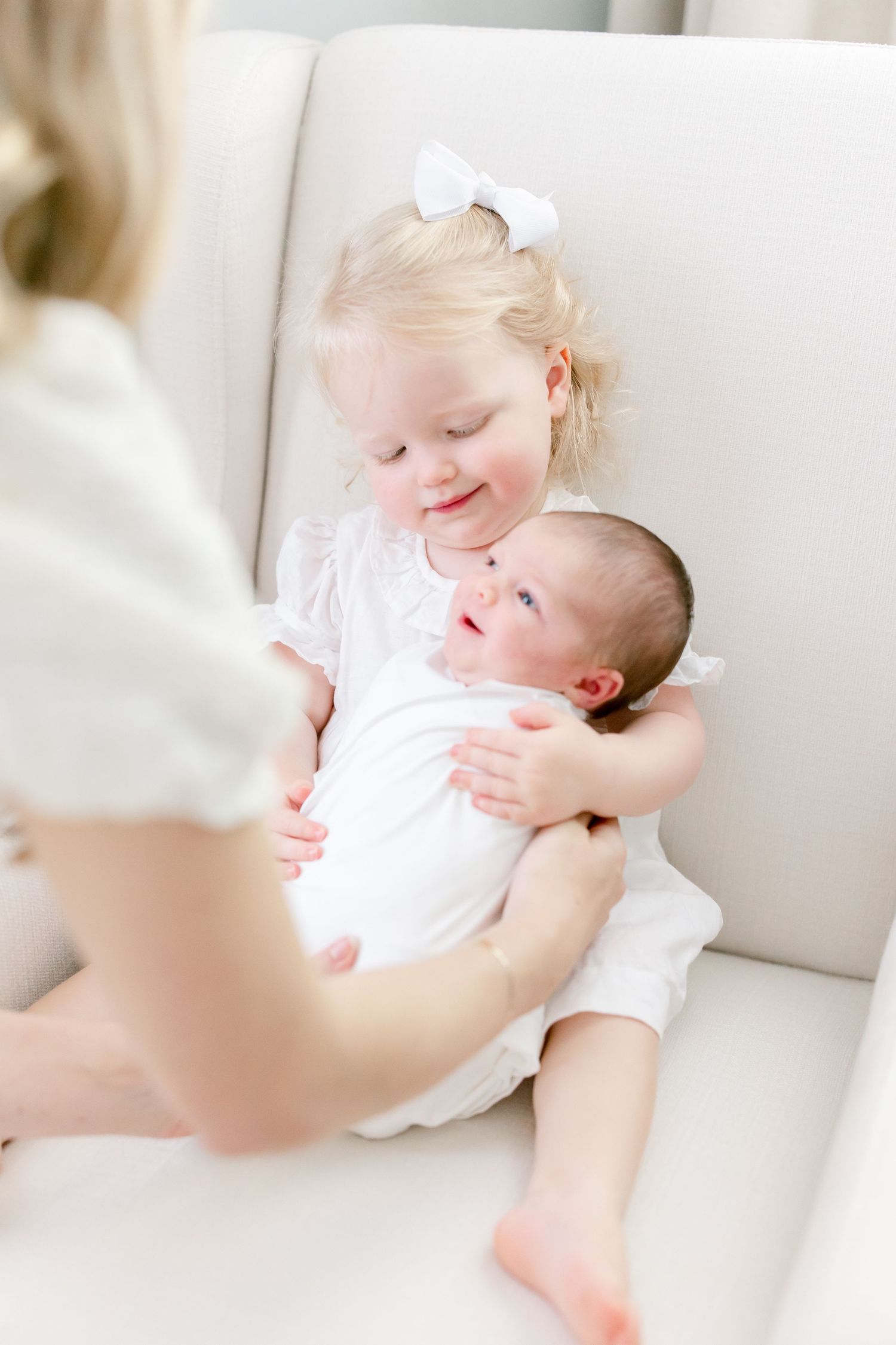 Two young children in white clothing share a sweet moment while sitting together on a white chair.