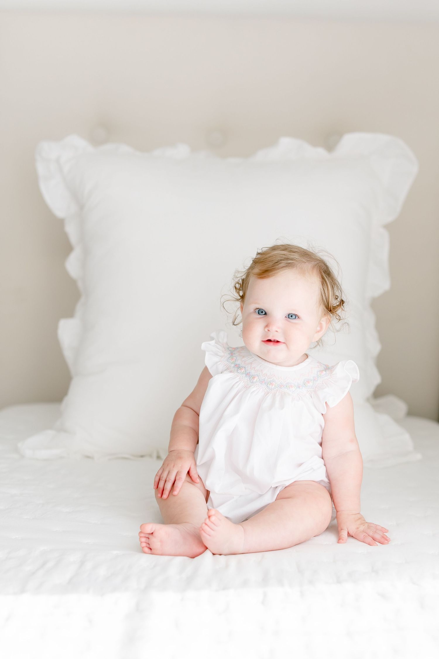 A baby wearing a white ruffled romper sits on a bed with white pillows and linens.