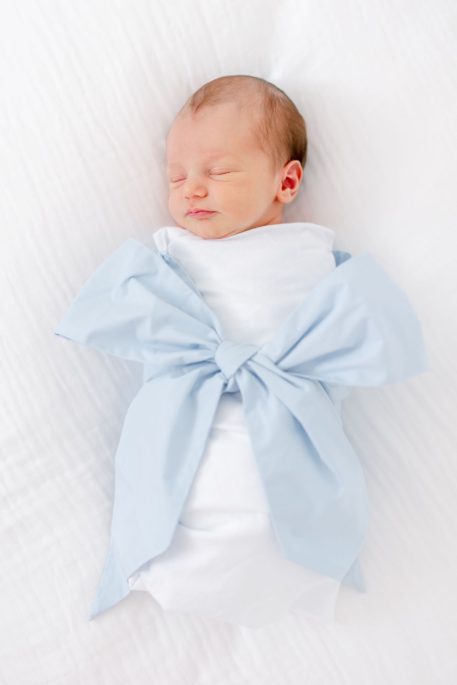 A newborn baby sleeping peacefully wrapped in white and light blue blankets tied in a bow.