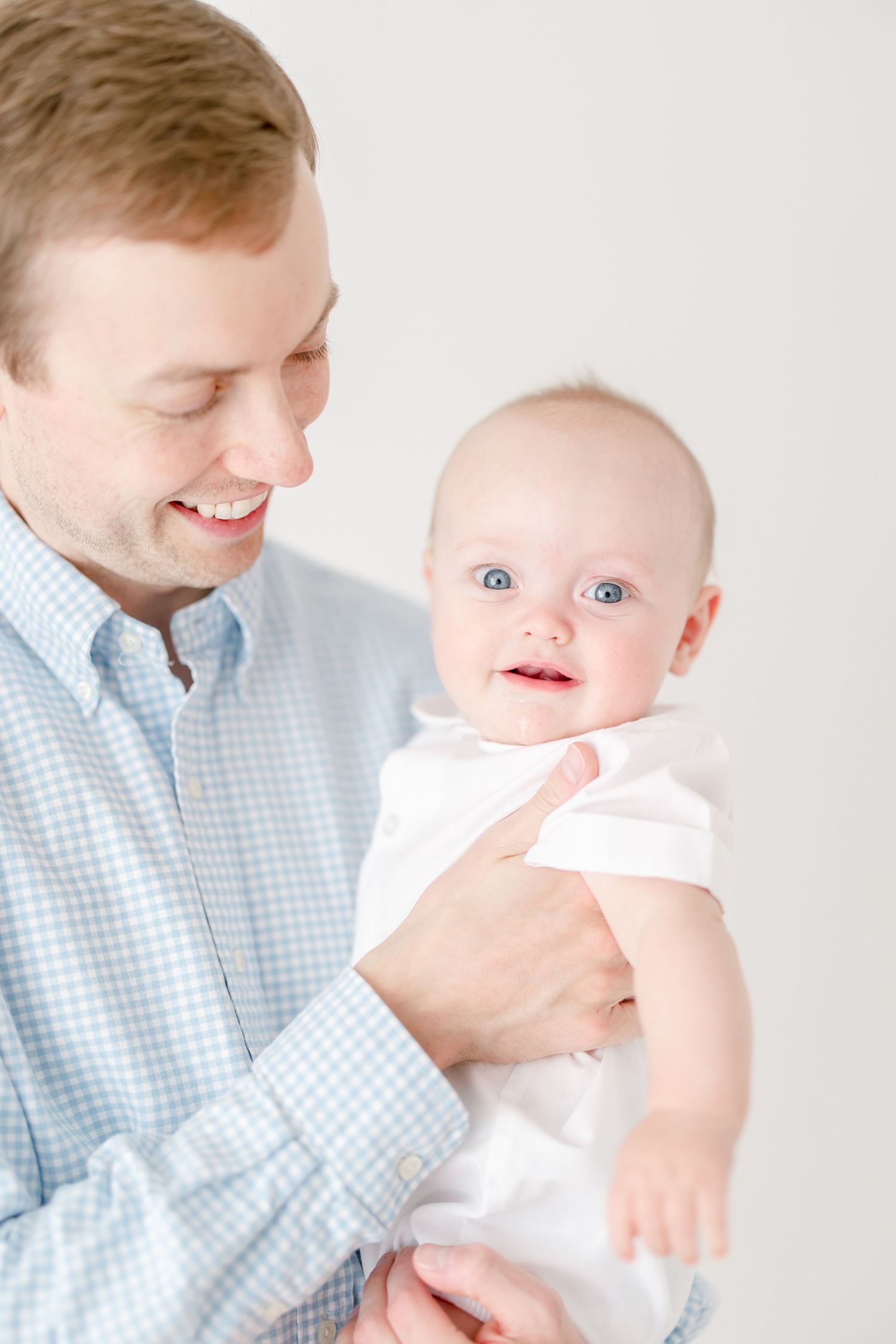 Parent in blue checkered shirt holding smiling baby in white outfit against white background.