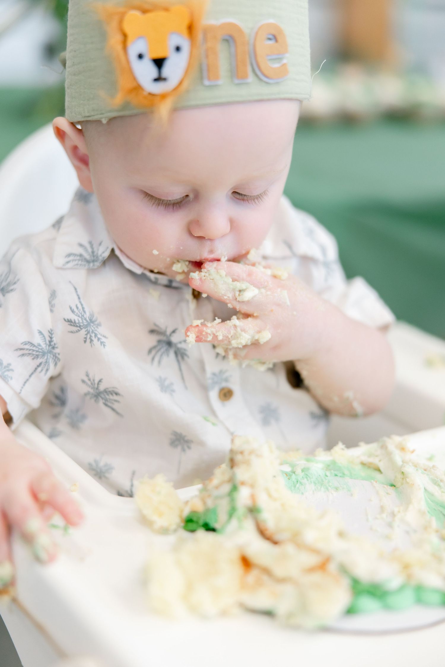 Baby in high chair wearing crown with fox design while eating birthday cake.