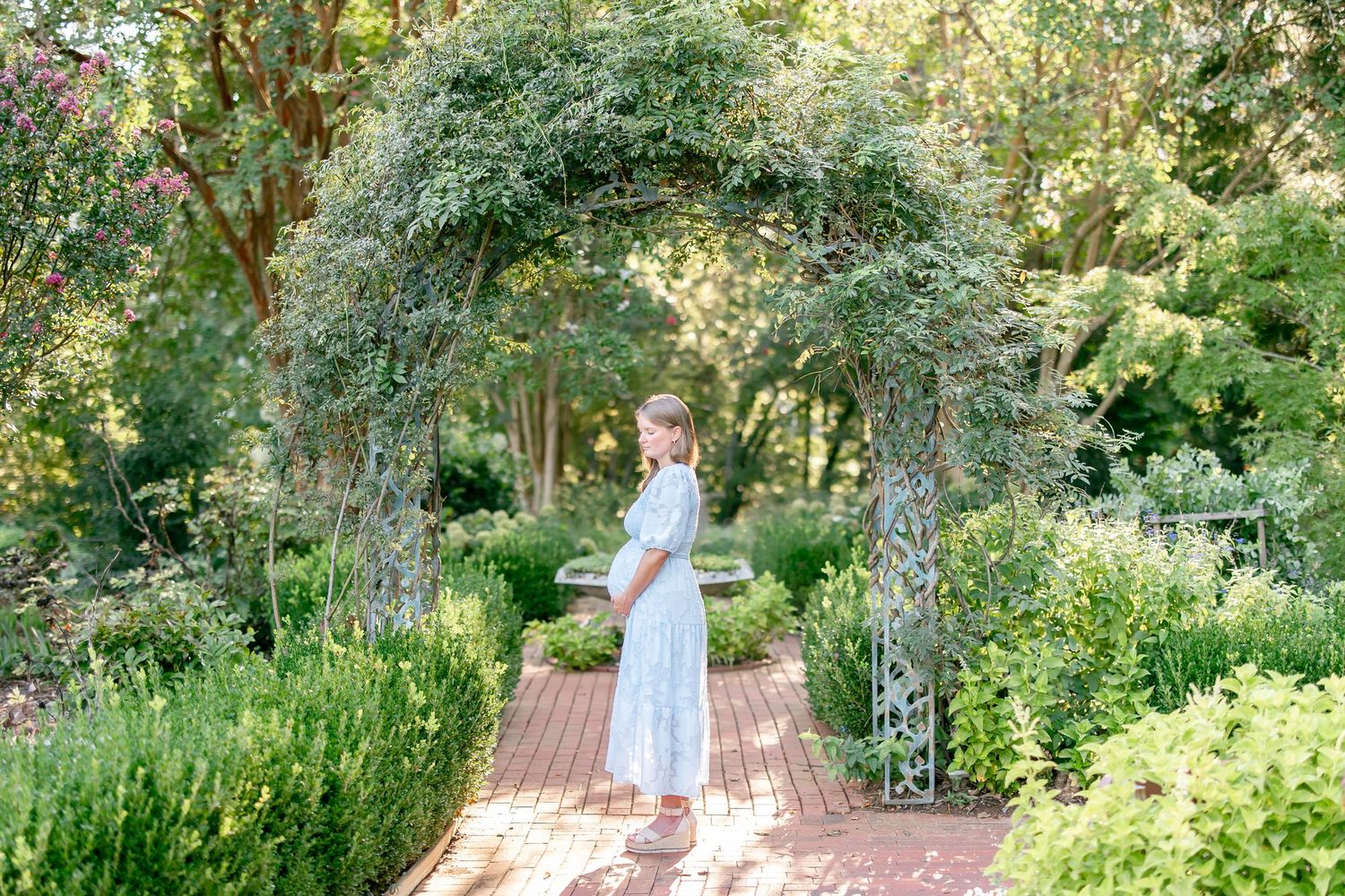 A figure twirls in a light blue dress on a wooden platform surrounded by lush green garden foliage on a sunny day.