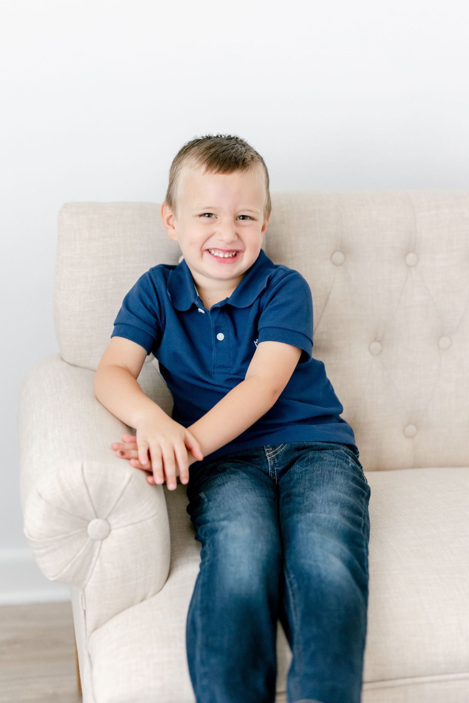 Young boy in navy polo sits comfortably on light beige couch showing joyful expressions.