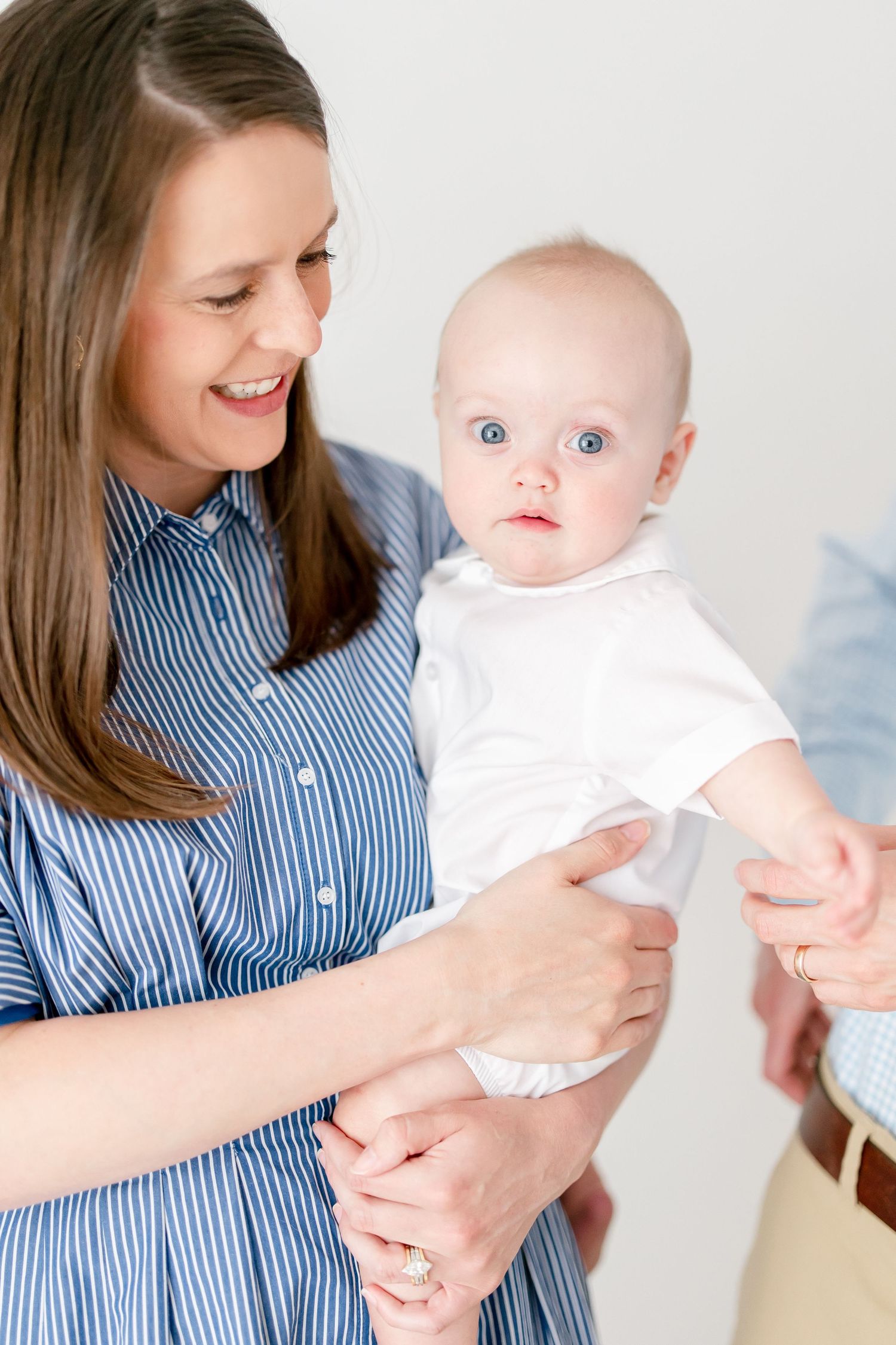 Baby in white outfit being held against blue and white striped shirt in bright minimal setting.