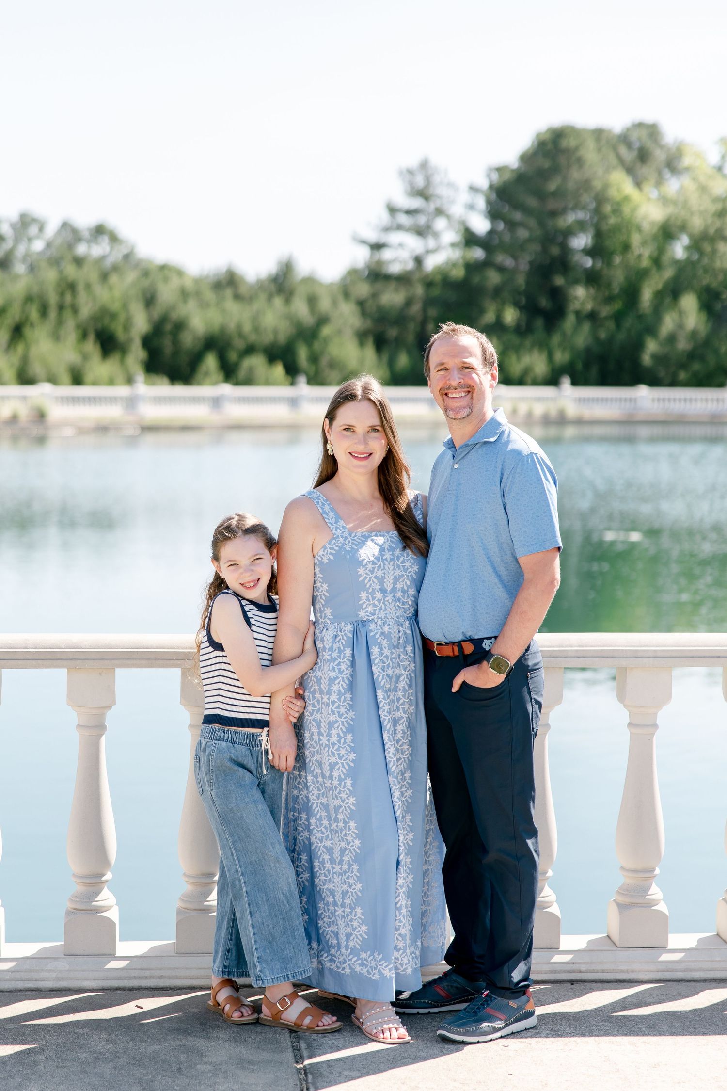 Family wearing coordinated blue outfits poses together on white balcony overlooking peaceful lake.