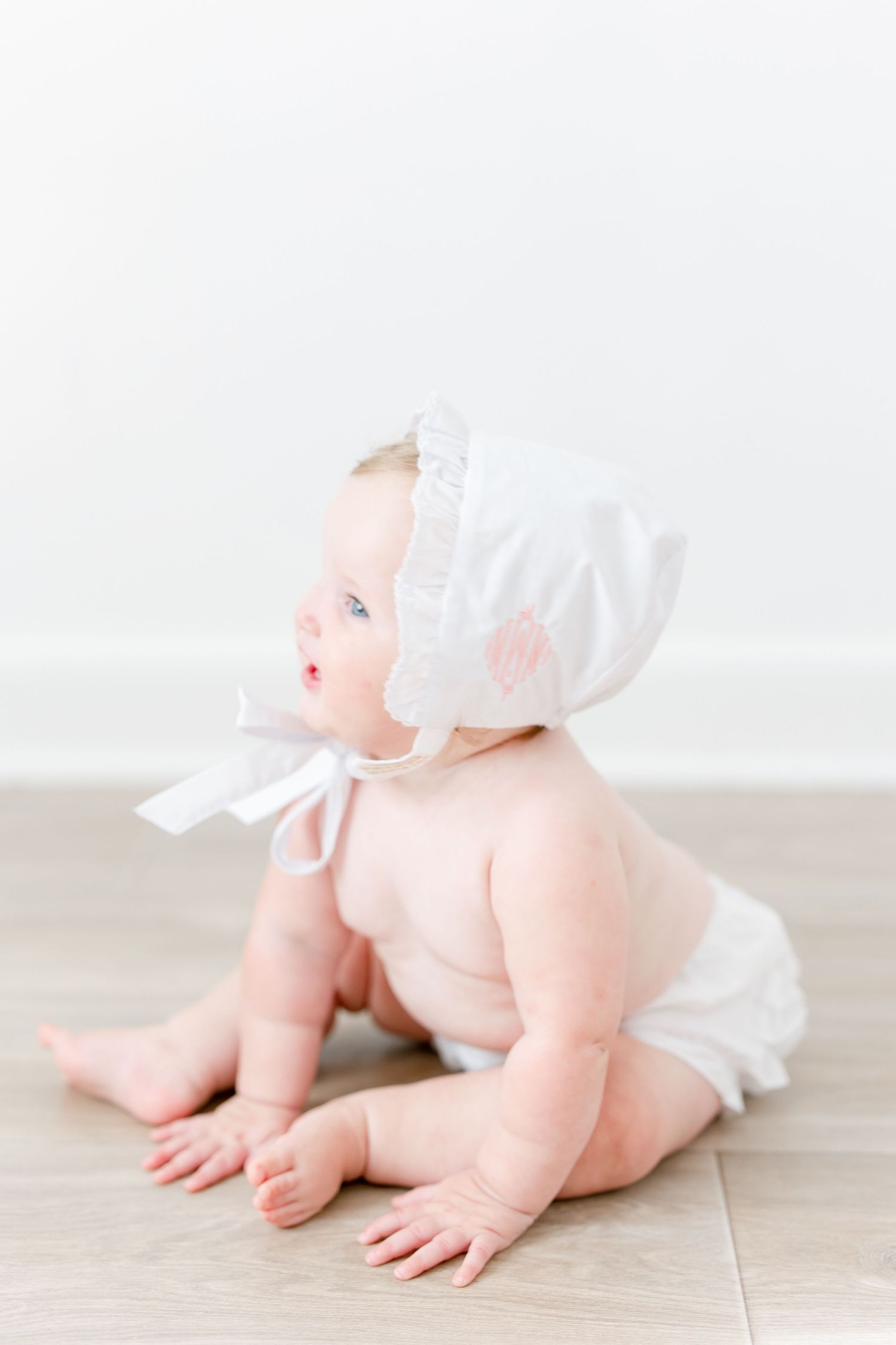 Sweet baby dressed in white bonnet and bloomers sitting on light wood floor in minimalist portrait setting.