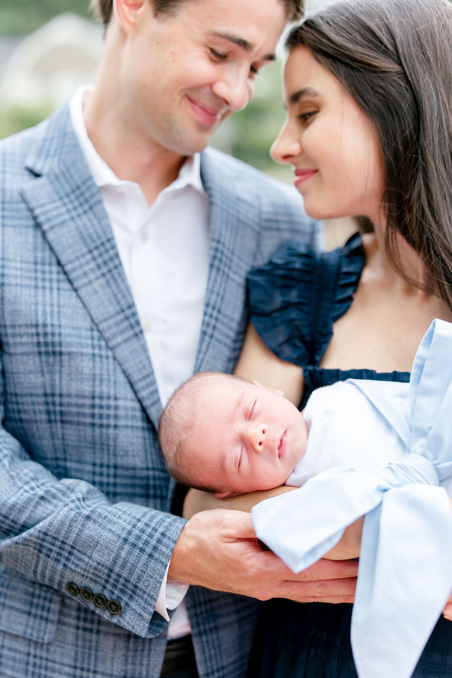Newborn photography session with parents in plaid blazer and navy dress holding infant.