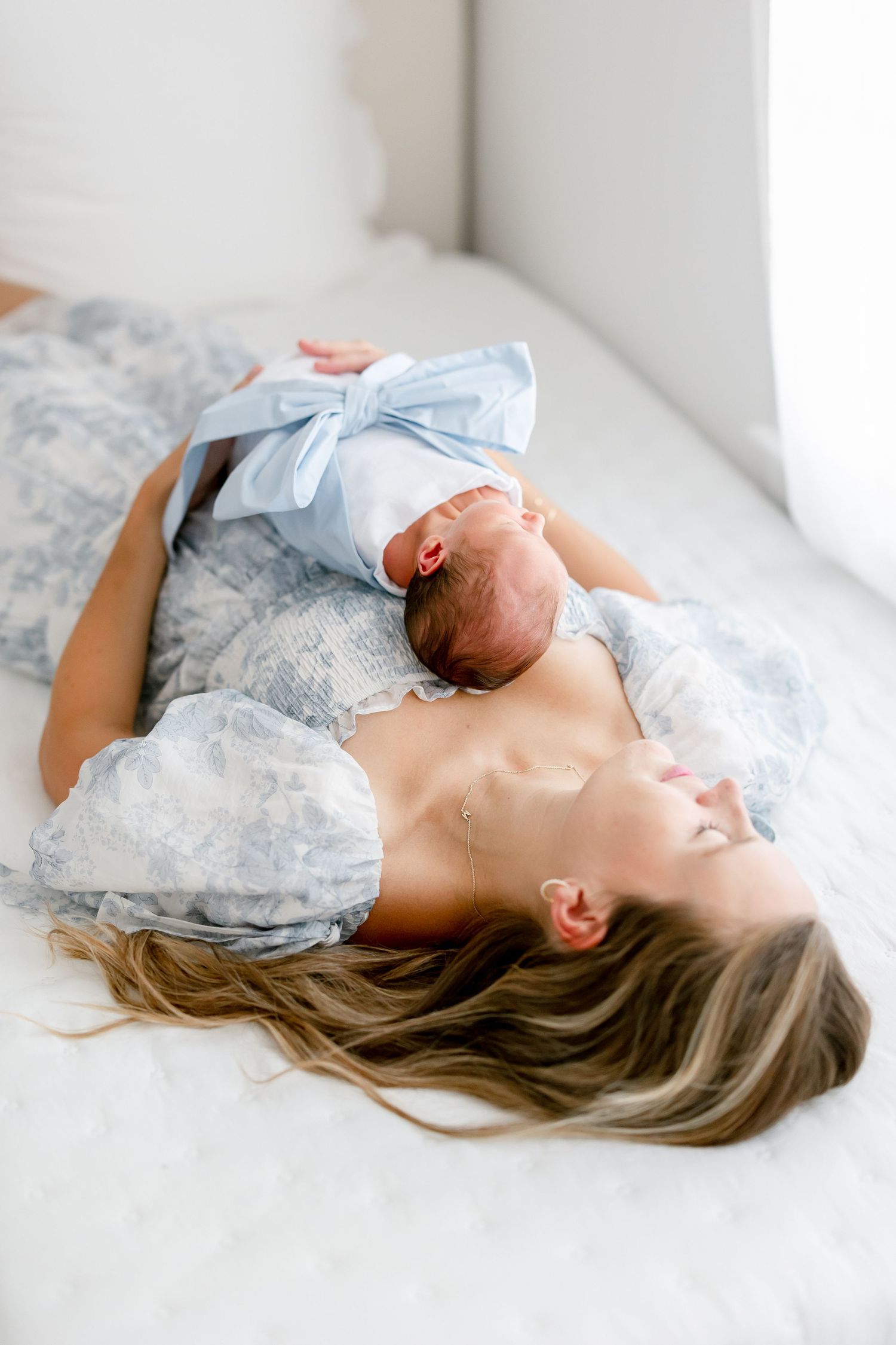 Mother and newborn baby wearing matching pale blue outfits laying together peacefully on white bedding.