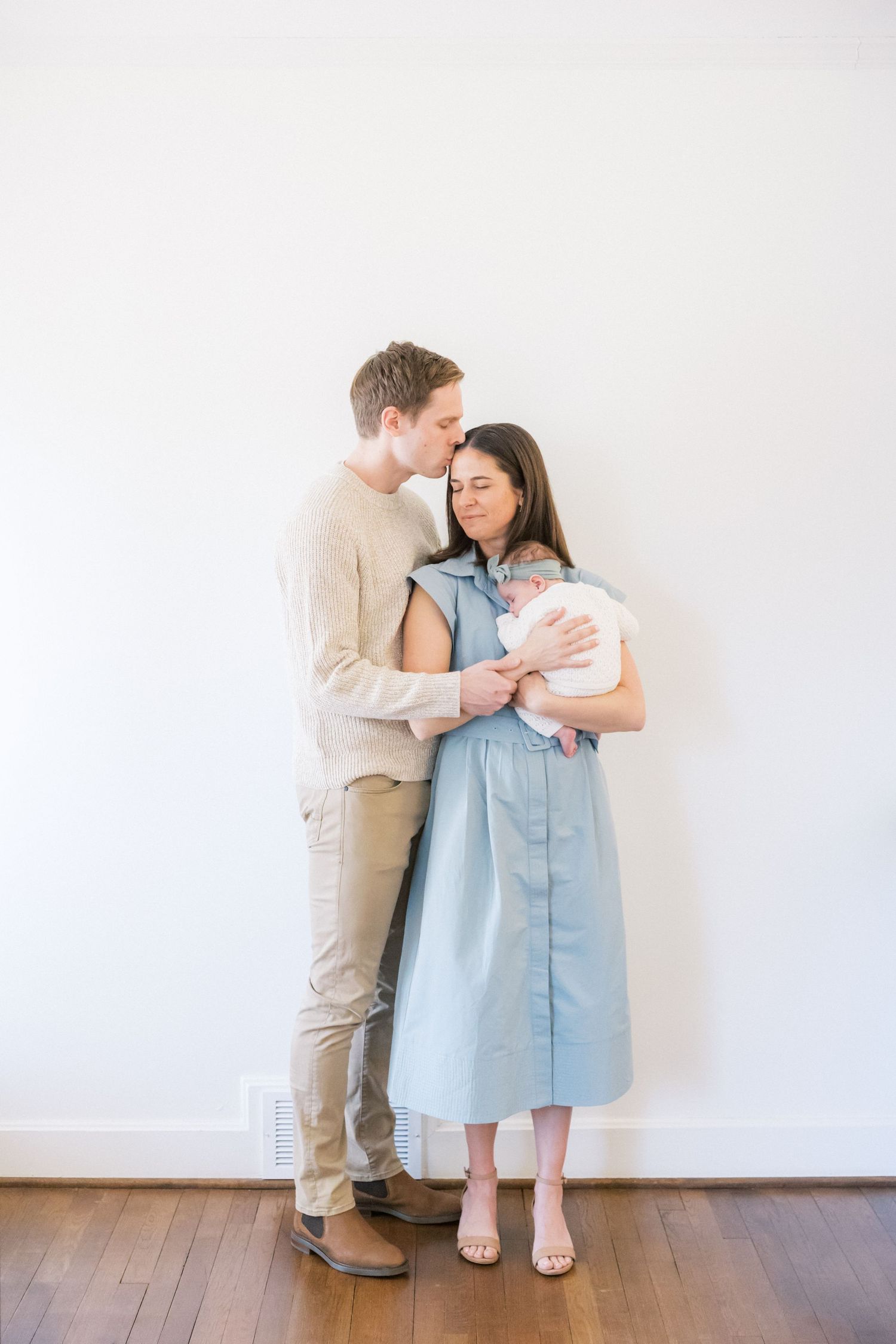 Family standing against white wall sharing intimate moments during newborn photoshoot.
